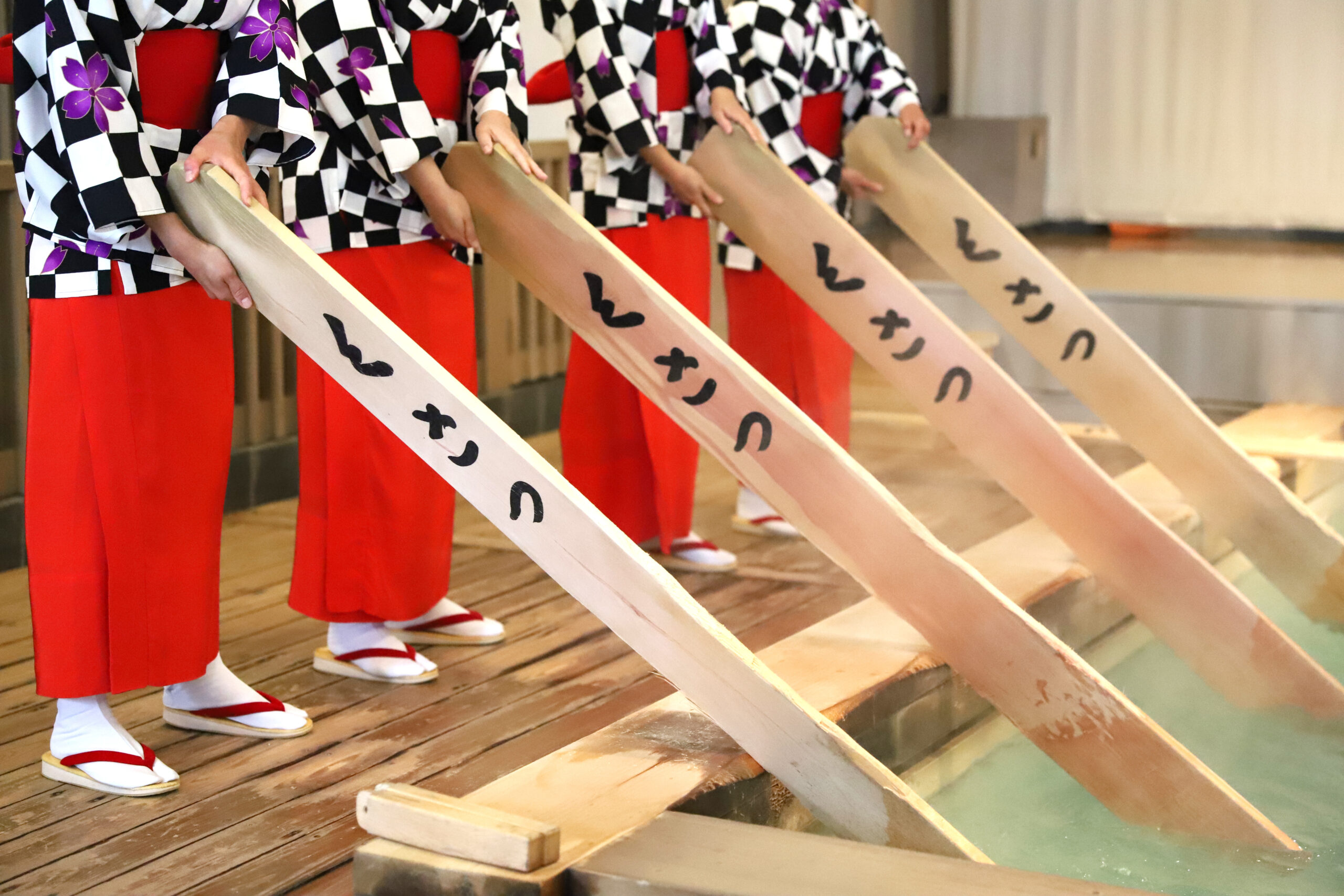 Group of women in black-and-white checkered kimonos with red sashes, holding long wooden boards with writing, standing on a wooden deck by water.
