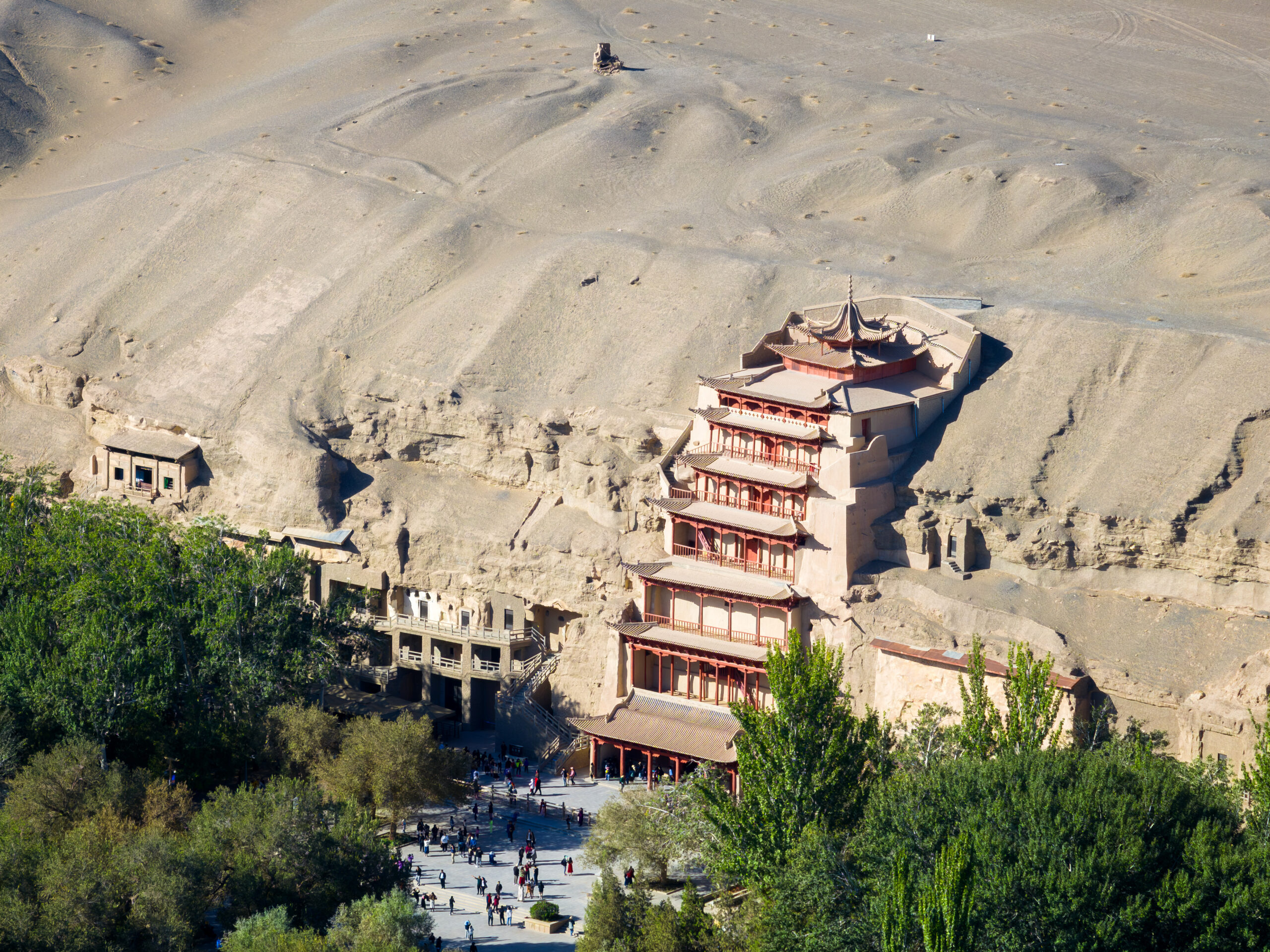 Tiered pagoda-style temple built into a rocky desert cliff, with visitors strolling along a plaza at the base and trees in the foreground.