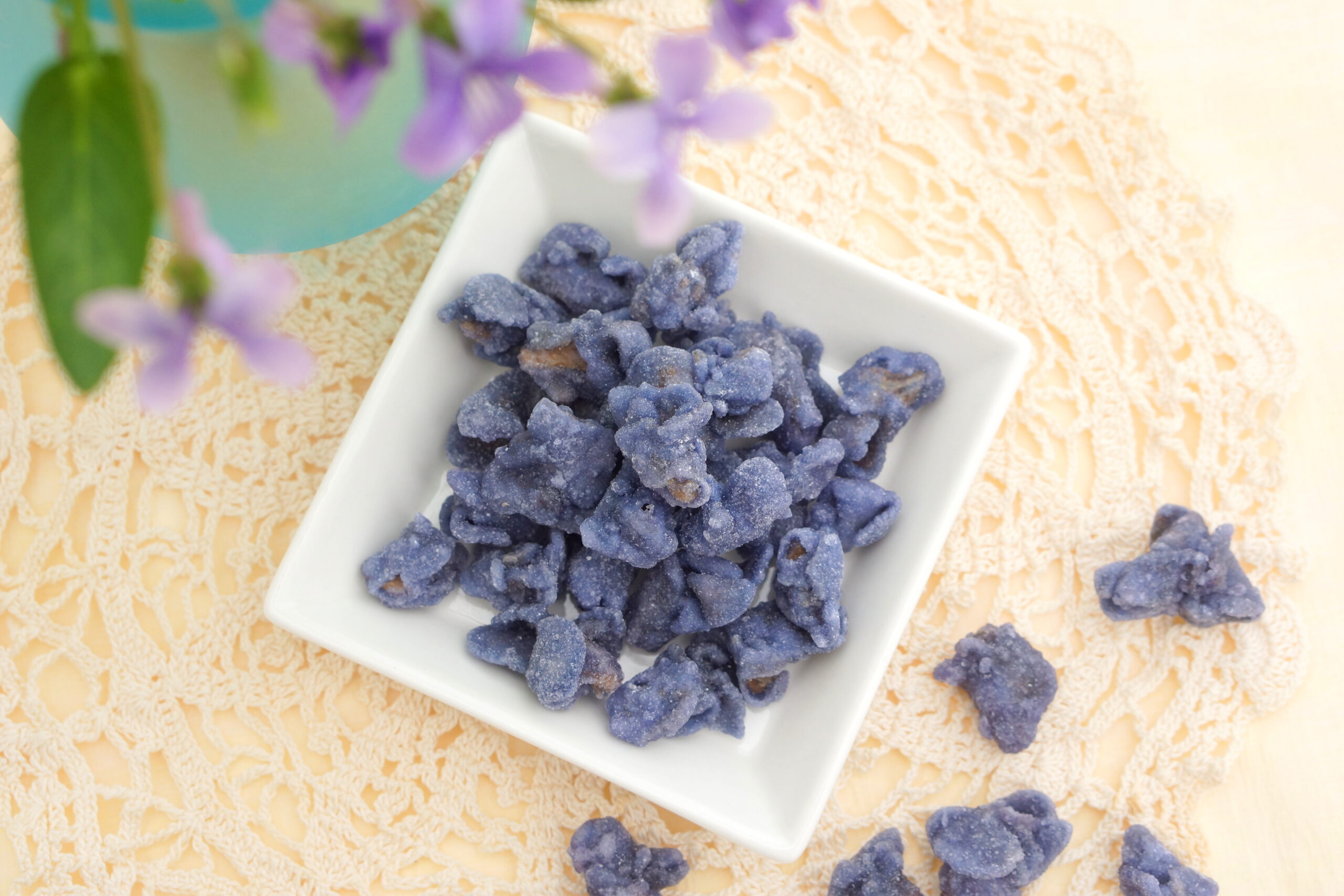 White square dish filled with blue-purple sugar-coated candies on a cream lace tablecloth, with a teal vase and purple flowers in the background.