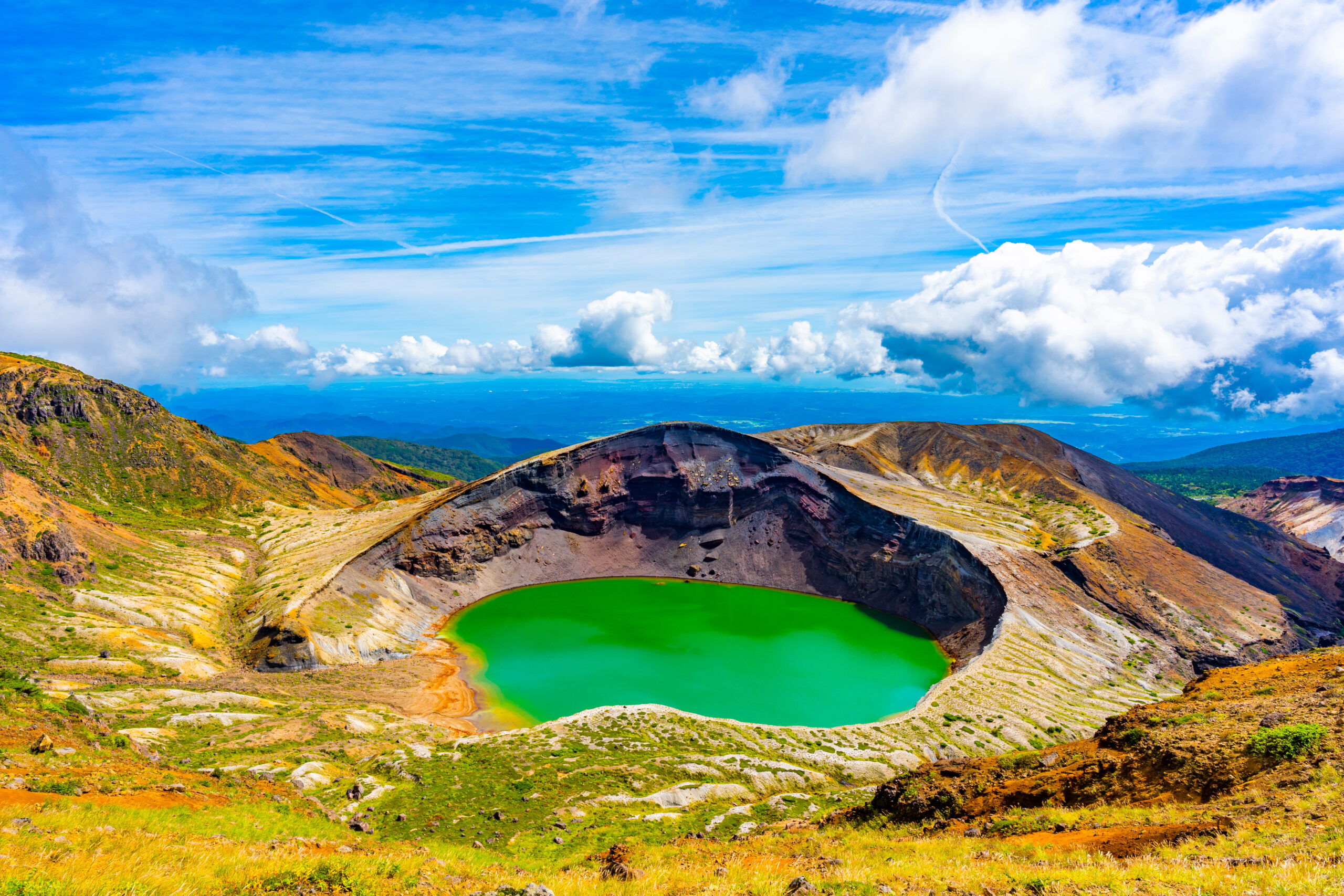 Bright green crater lake surrounded by barren volcanic hills under a blue sky with clouds.