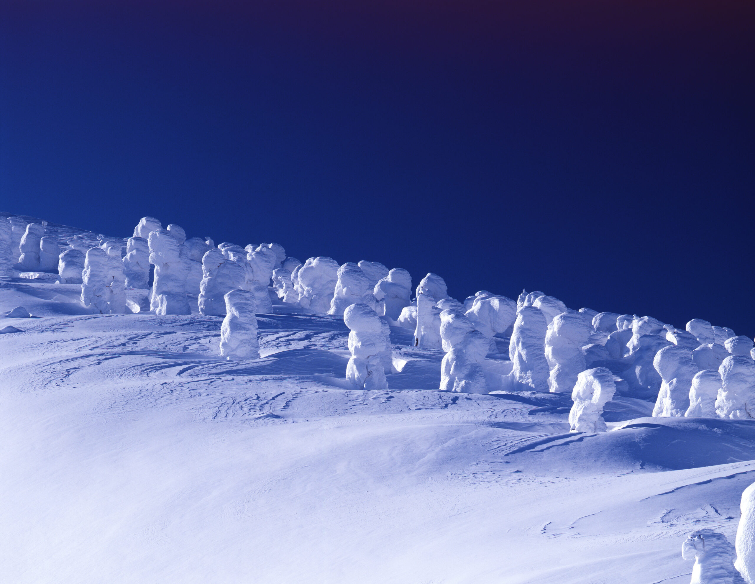 Snow-covered landscape with clustered ice formations along a slope under a deep blue sky at dusk (or dawn).