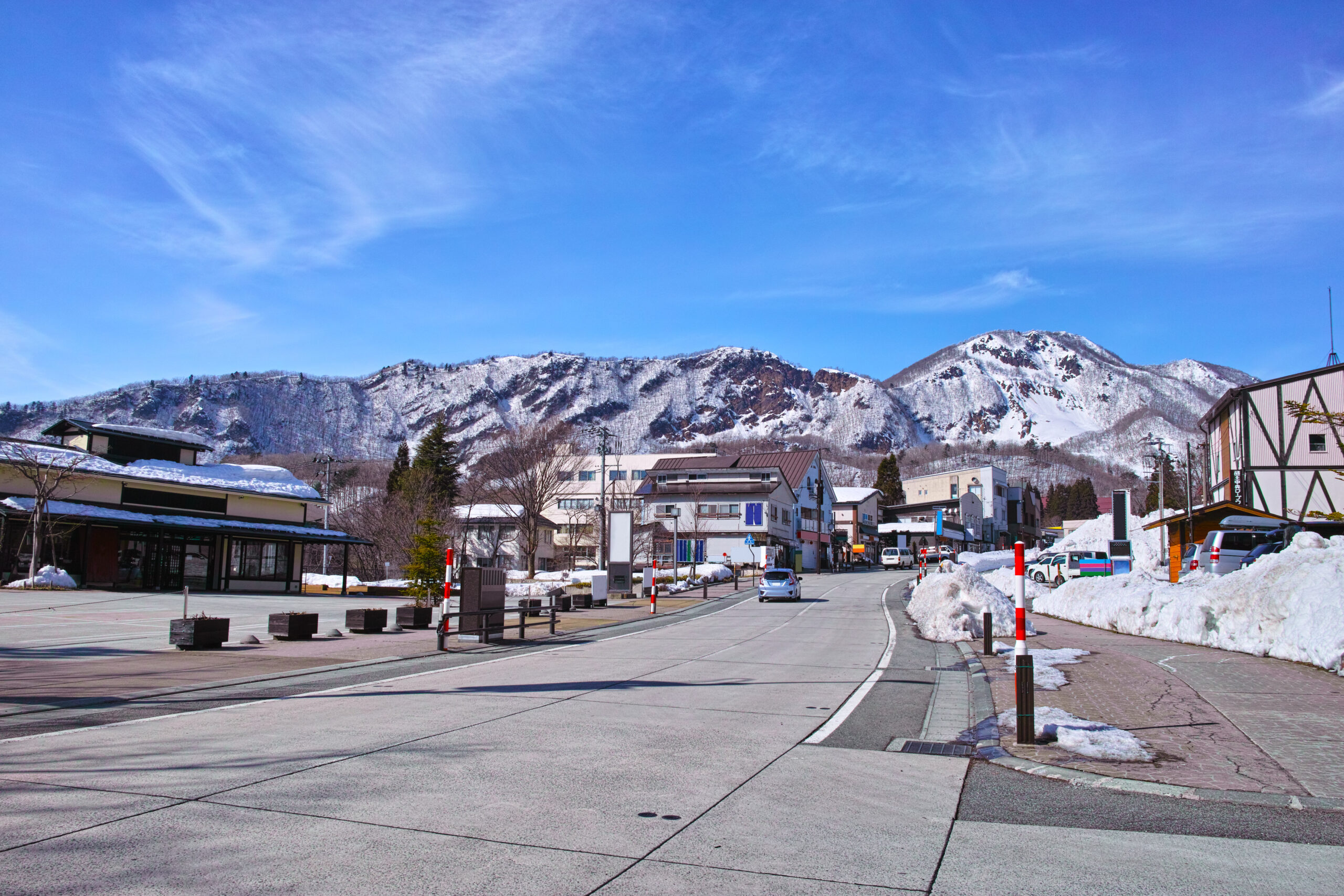 Snowy town street with buildings and parked cars, mountains in the background under a blue sky.