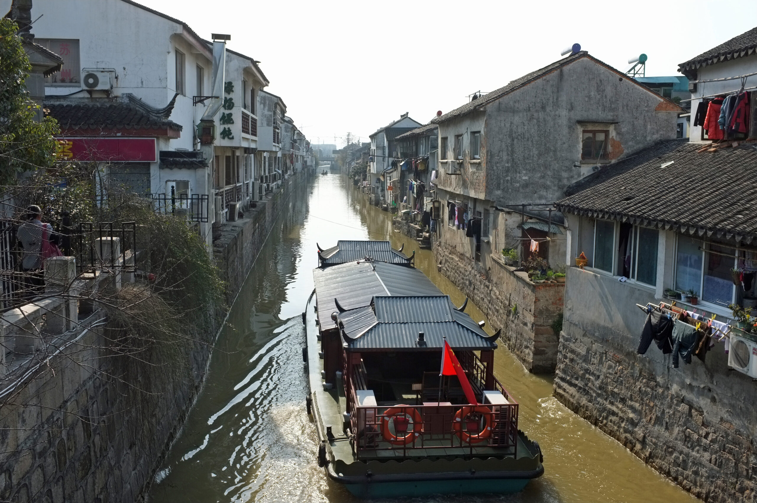 Narrow canal flanked by old white buildings with tiled roofs; a traditional boat with a red flag travels down the waterway, clothes hang along the right wall.