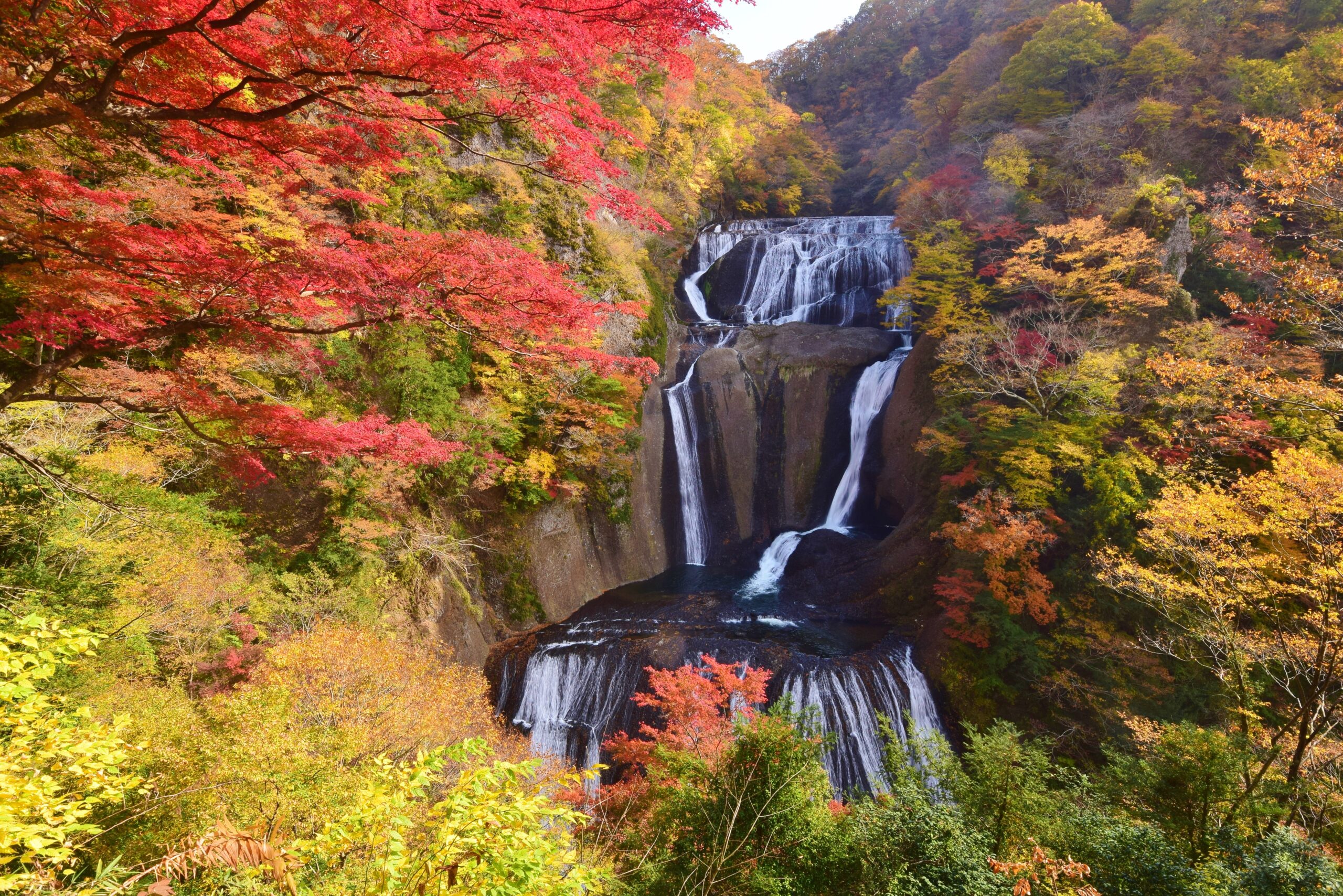 Autumn forest with red, yellow, and orange leaves surrounding a multi-tier waterfall cascading over rocky cliffs.