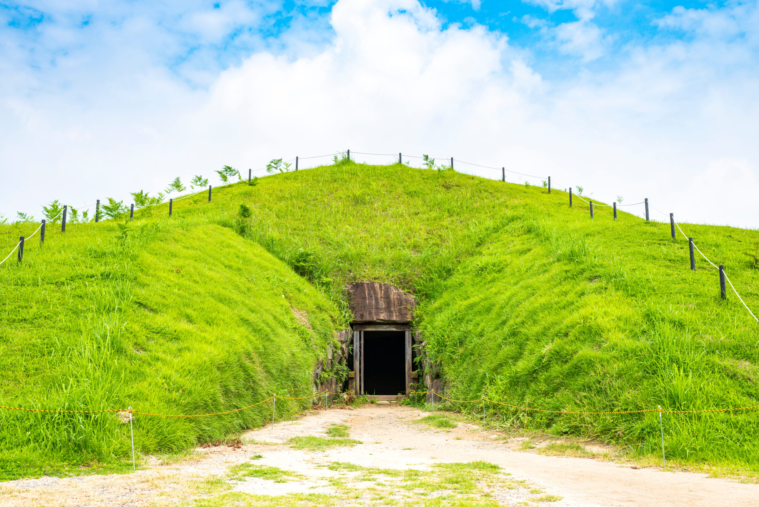 Grass-covered hill with a dark tunnel entrance at its base and a dirt path leading to it; rope barriers run along the sides.