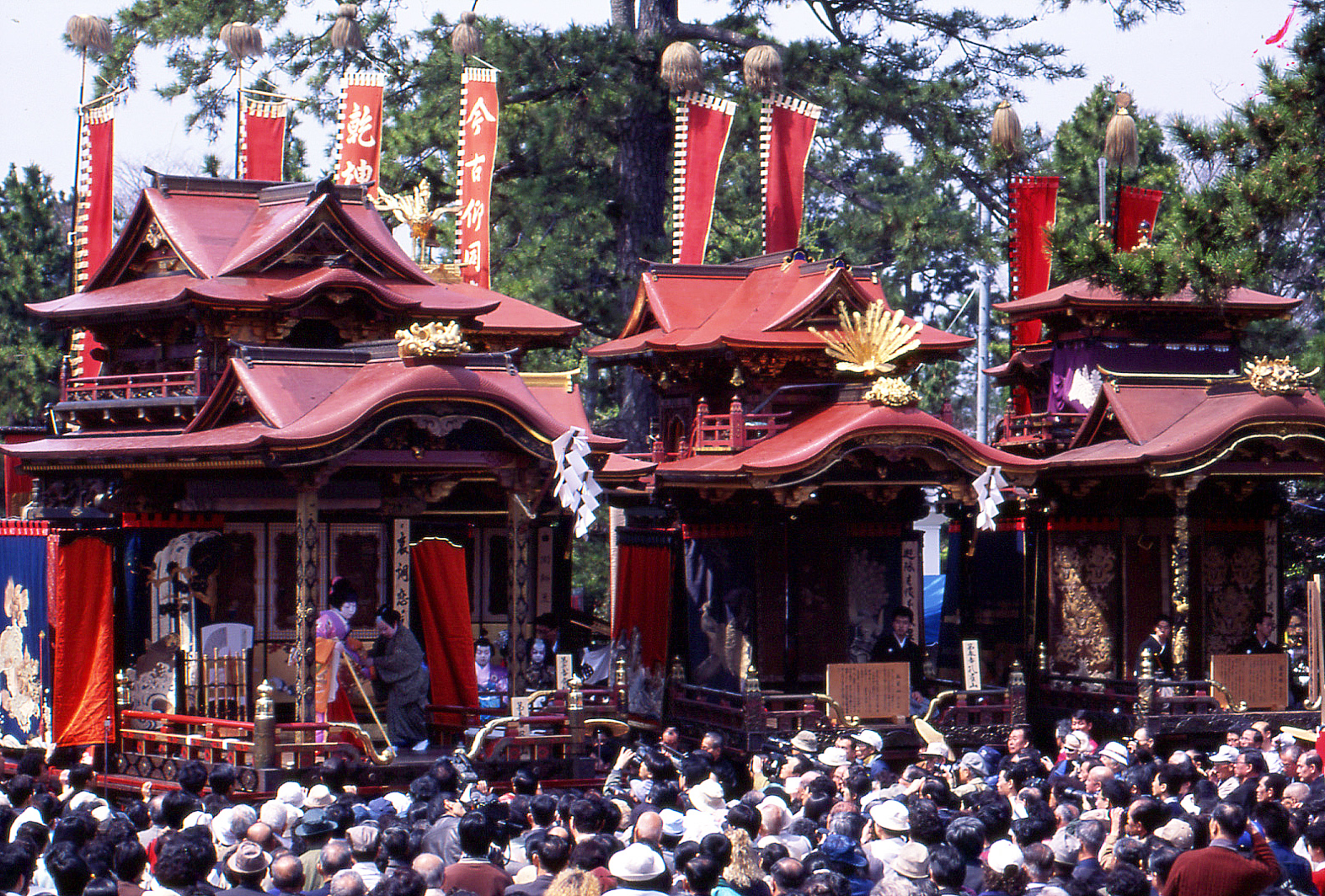Elaborate red festival floats with gold decorations passing through a crowded traditional Japanese matsuri scene.