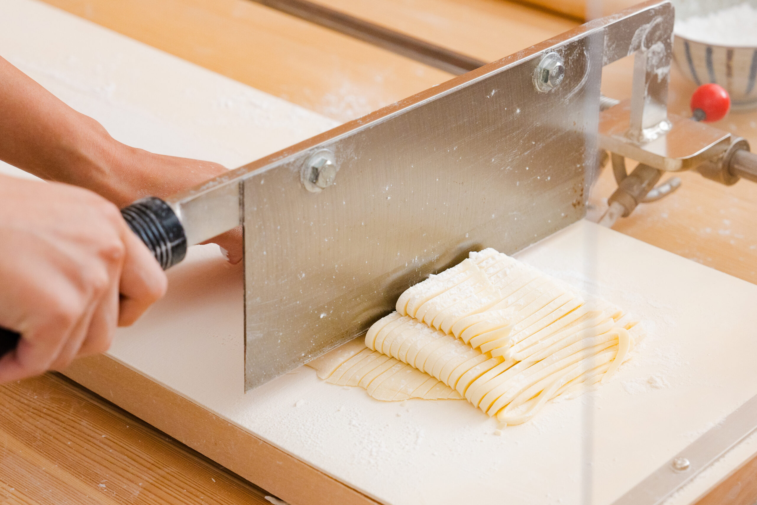 Person operates a metal pasta machine, pressing dough into long ribbon-like strips on a flour-dusted wooden surface