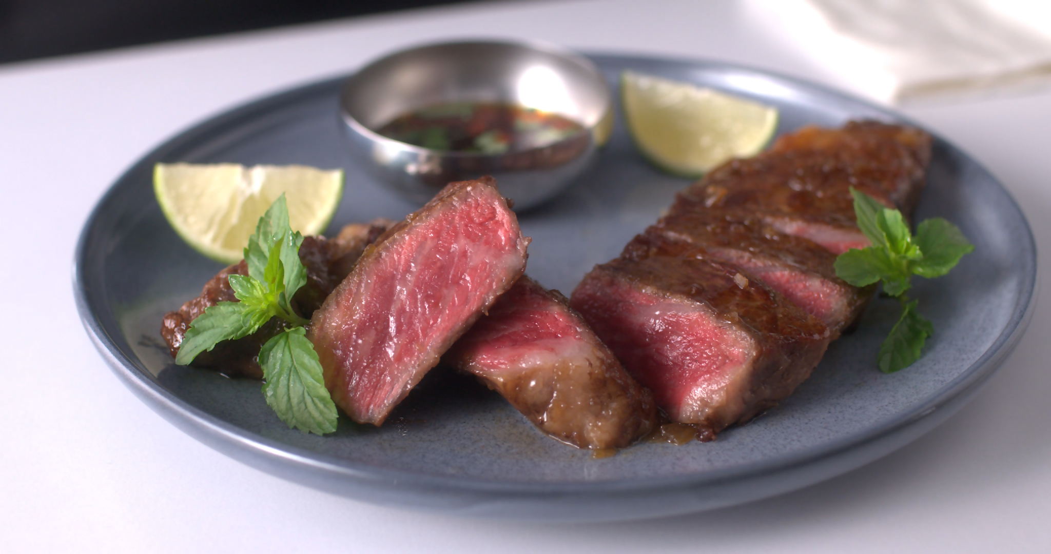 Plate of medium-rare steak slices with lime wedges and mint, plus a small metal cup of sauce on a blue plate.