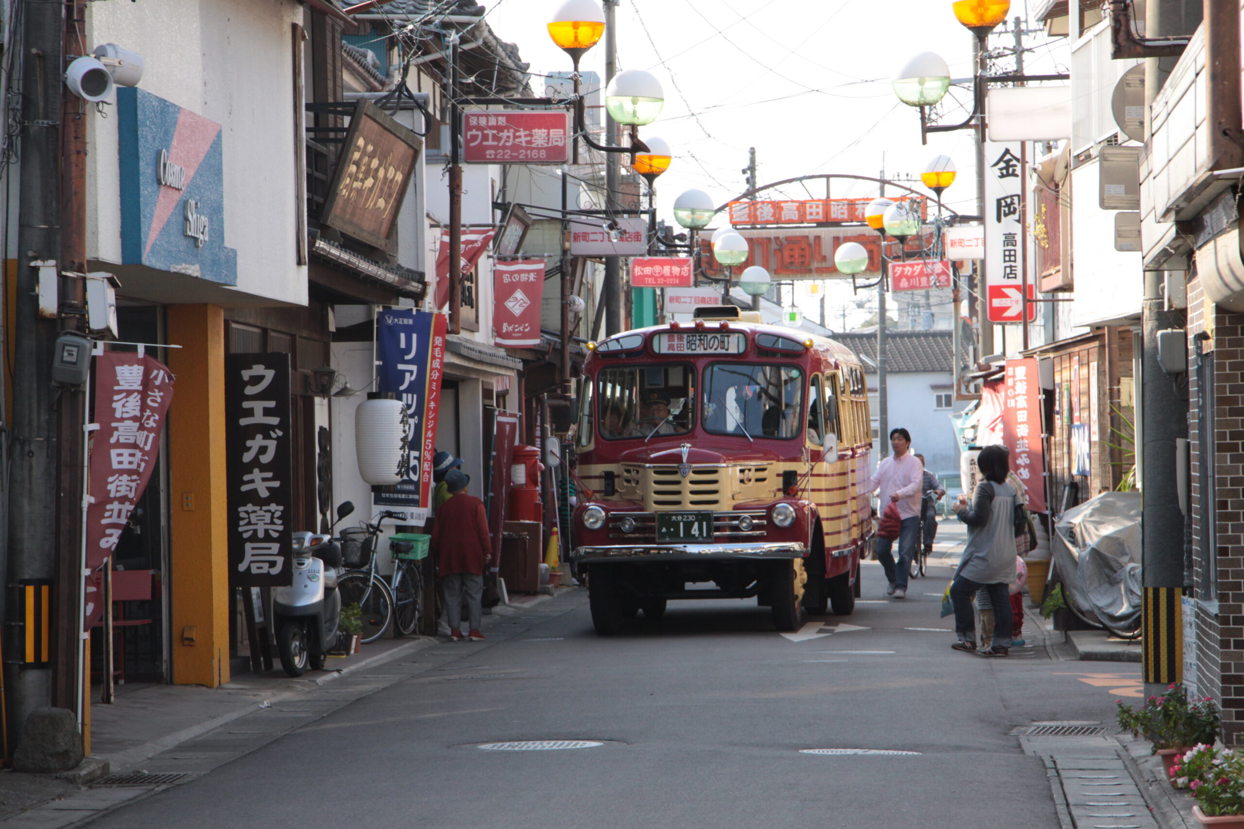Narrow Japanese street with a red vintage trolley bus in the center, storefronts and signs in Japanese lining both sides, pedestrians walking along.
