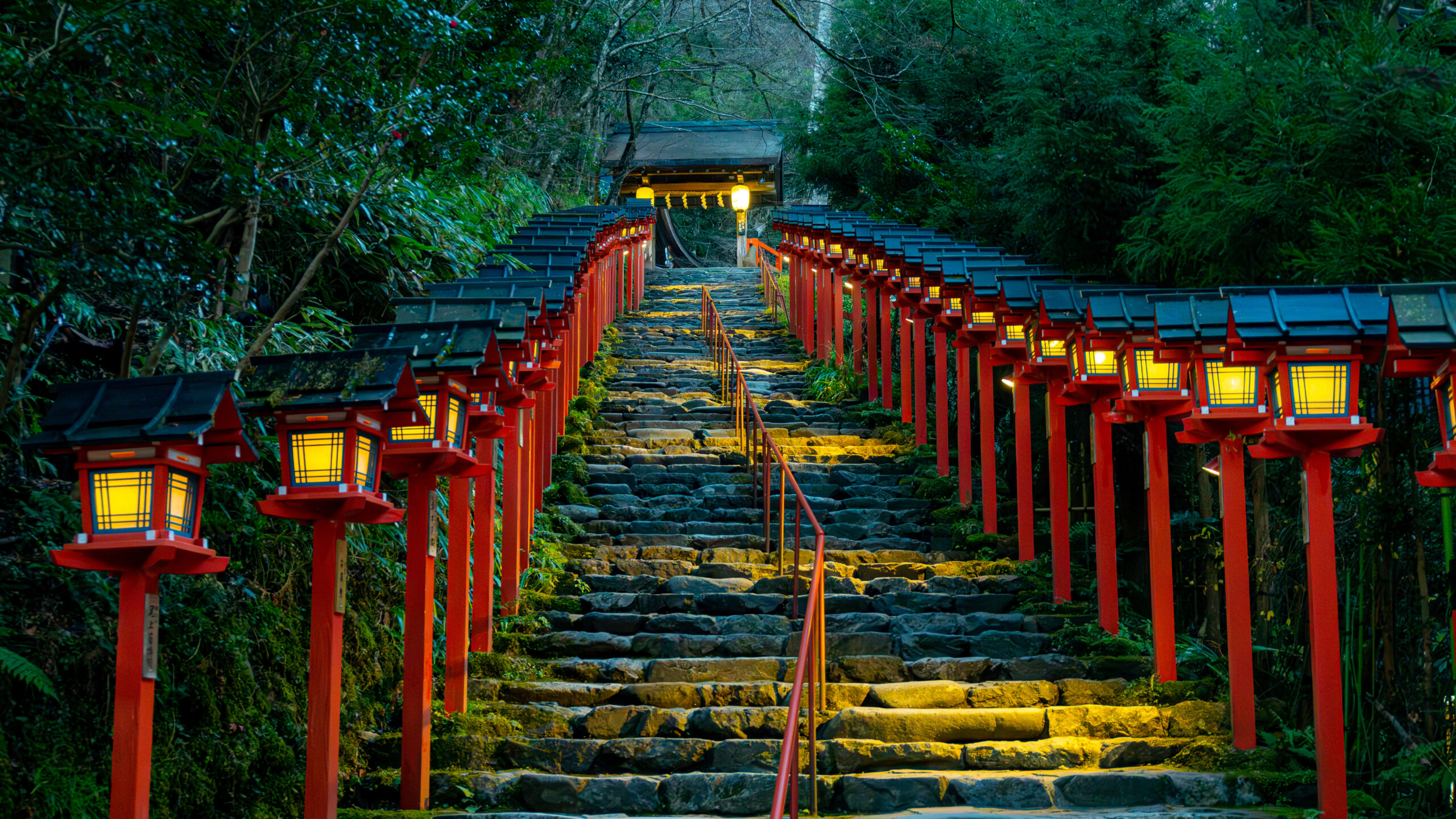 Stone steps lined with red lanterns lead through a lush forest toward a shrine entrance, glowing at dusk/dawn light.