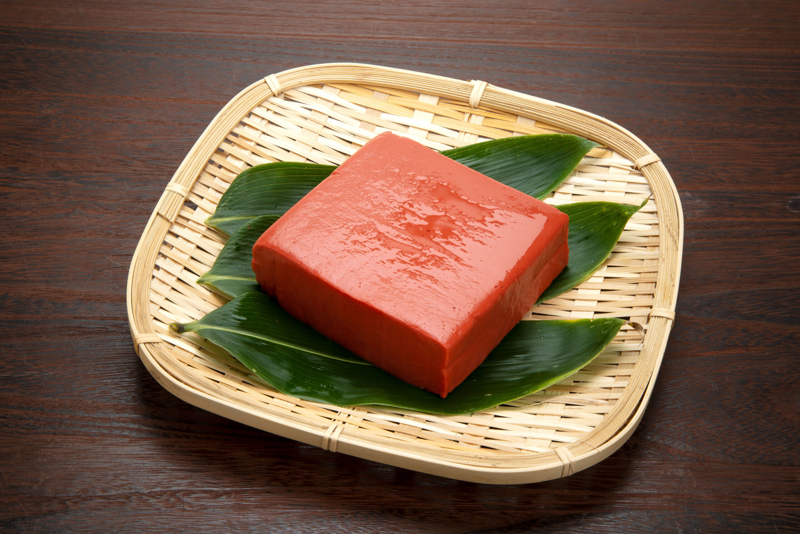 Red rectangular soap bar resting on green leaves inside a woven wicker basket on a dark wooden surface, close-up.