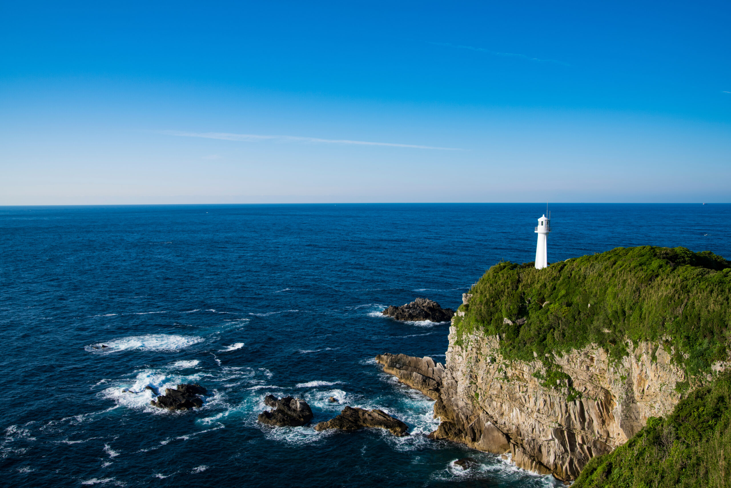 White lighthouse perched on a rocky cliff with green bushes overlooking the blue ocean and clear sky.