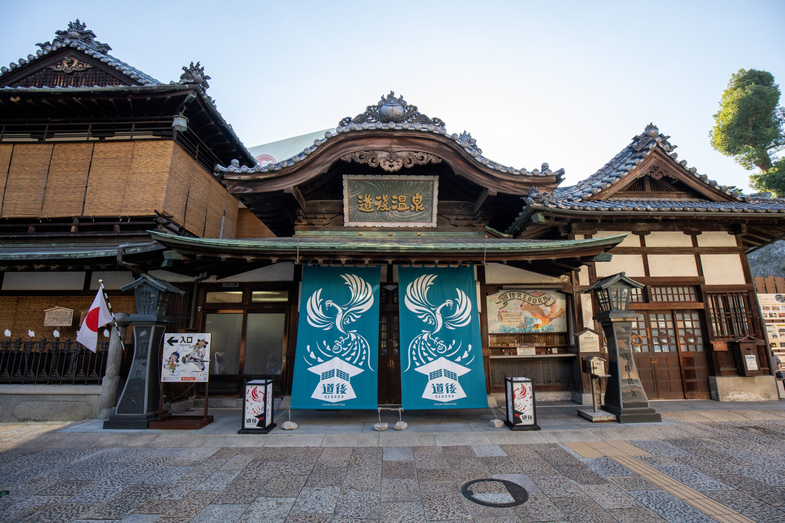 Traditional temple facade with ornate tiled roofs and carved wooden details, flanked by blue banners featuring white phoenix designs at the entrance.