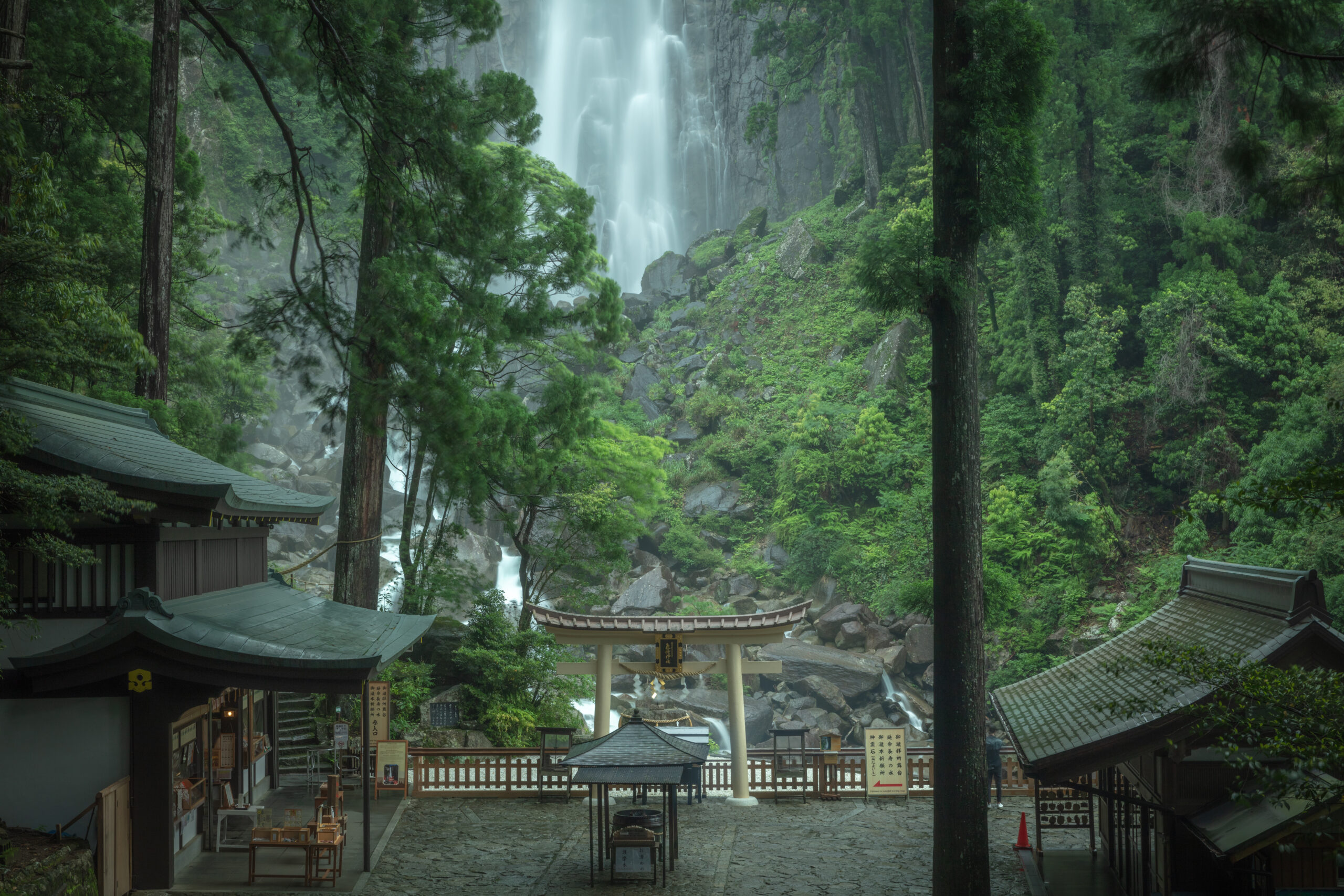 Temple courtyard with wooden buildings and a torii gate, surrounded by lush green forest and a distant waterfall in the misty background.