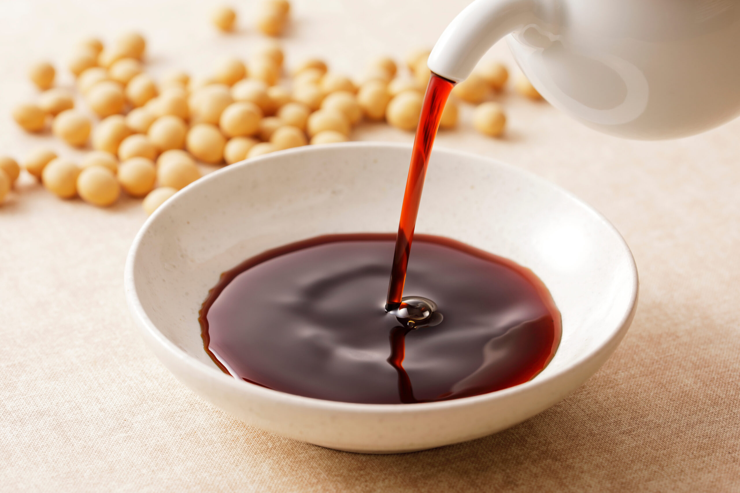 Pouring dark red sauce into a white bowl with soybeans scattered on a beige surface in the background, close-up.