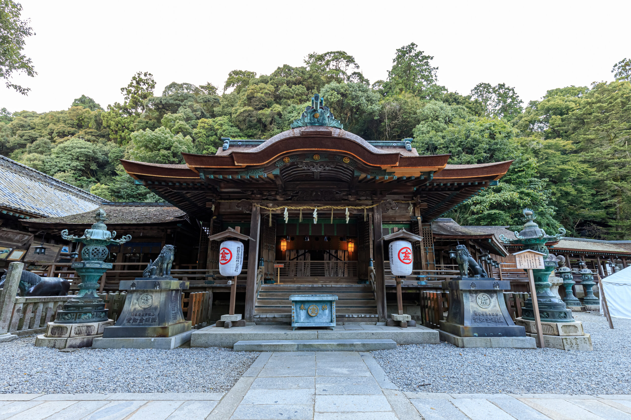 Front view of a traditional Japanese shrine with a curved, ornate roof, lanterns, and stone basins, set against a dense green forest backdrop.