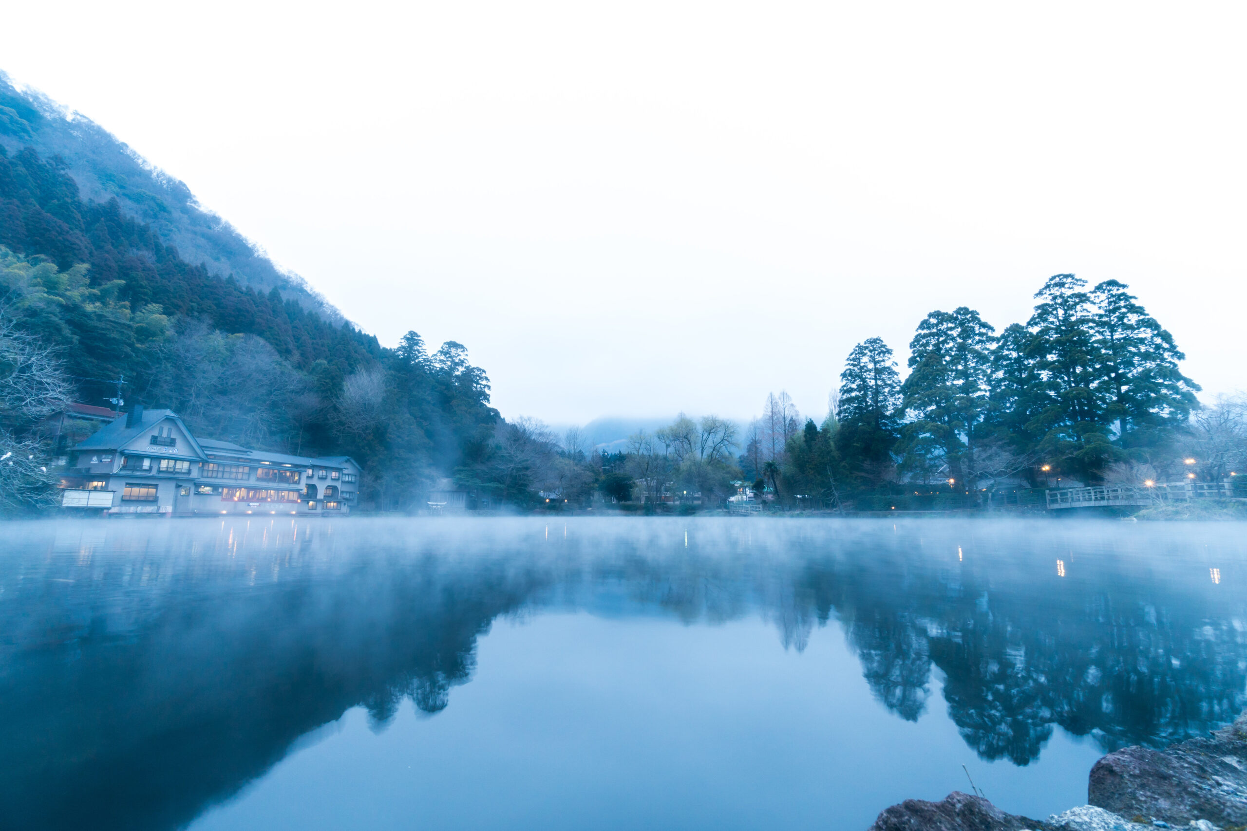 Misty lake scene with forested hills on the left, tall trees on the right, and a building along the shore reflected in calm water.