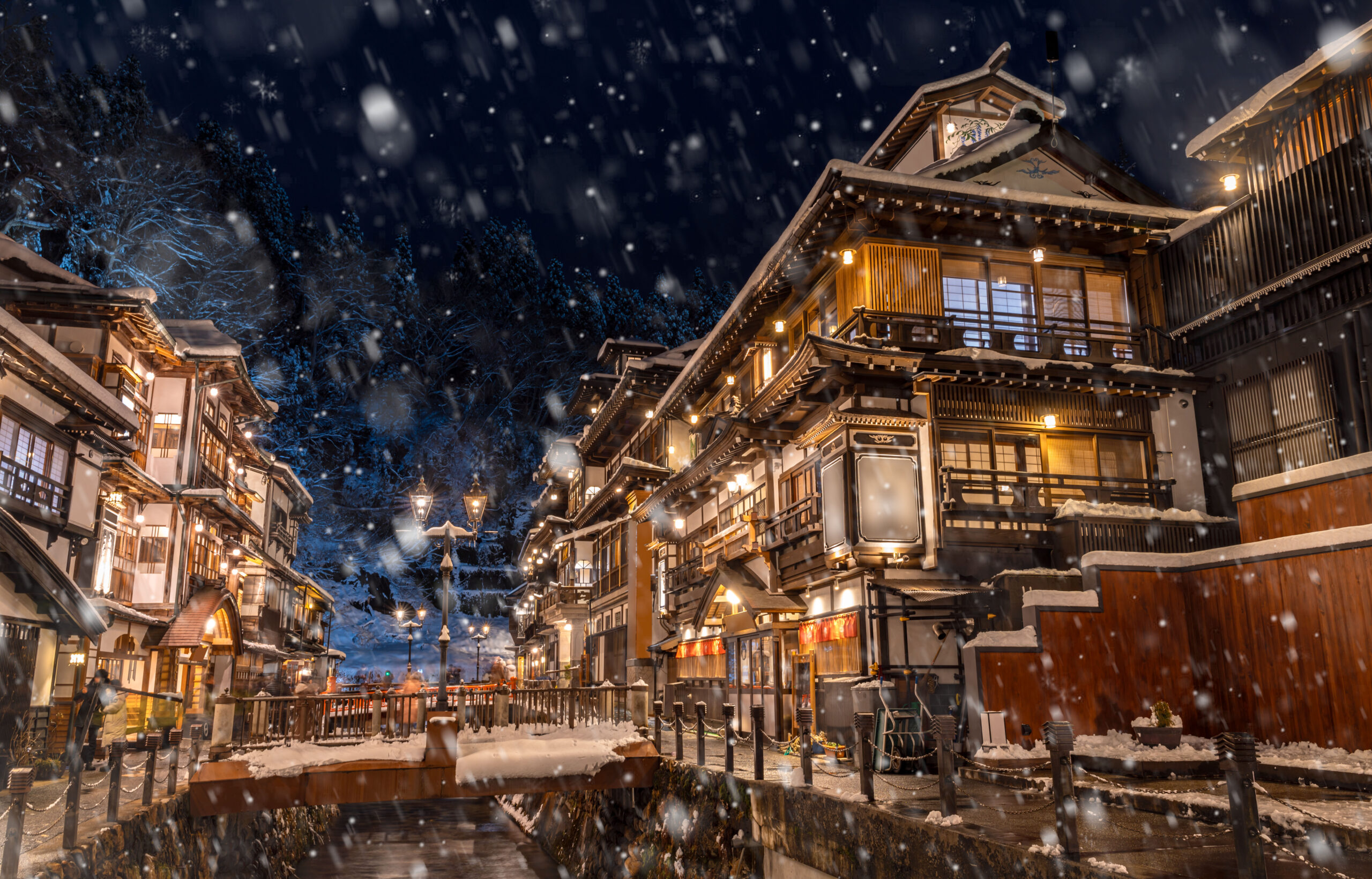 Snow falls over a traditional Japanese village at night, lanterns glow along snow-covered eaves and a canal in the foreground.