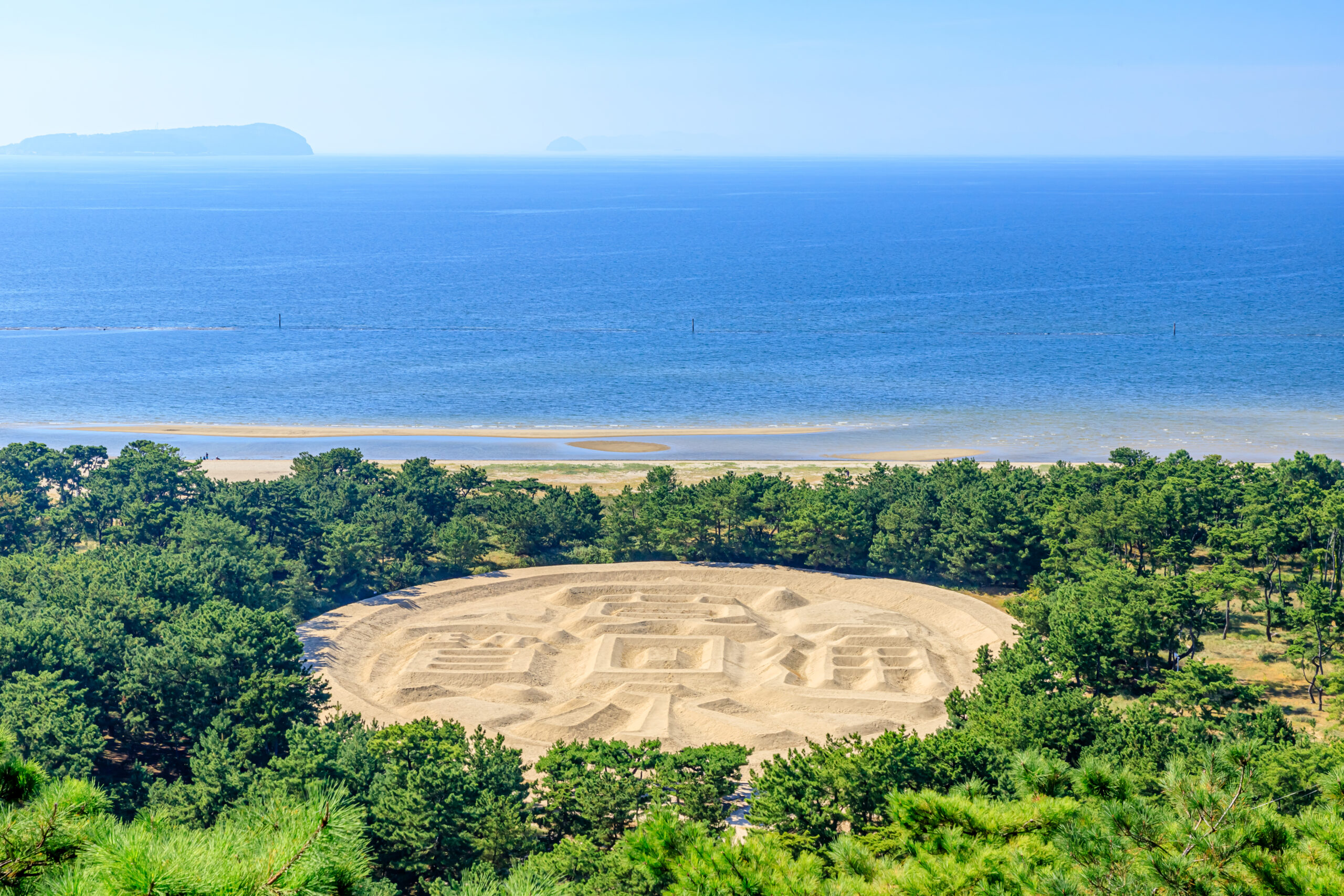 Dune field edged by pine trees overlooking a calm blue sea with distant islands on the horizon.