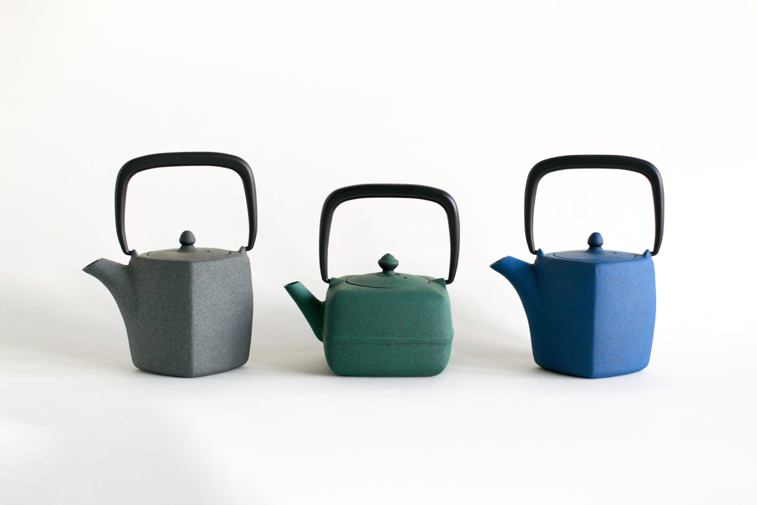Three matte kettles in gray, green, and blue with black handles lined up against a white background.