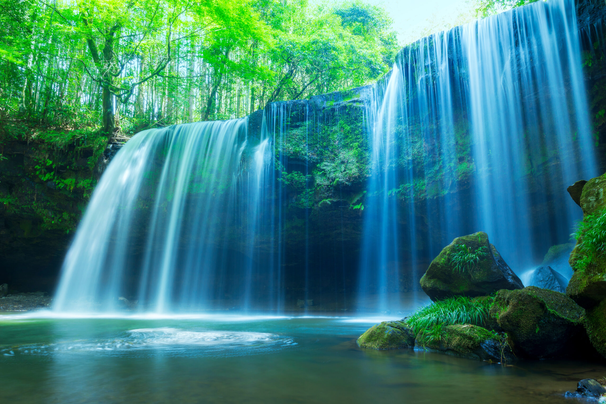 Waterfall cascading over a rocky ledge into a green, tranquil pool surrounded by lush trees and mossy rocks.