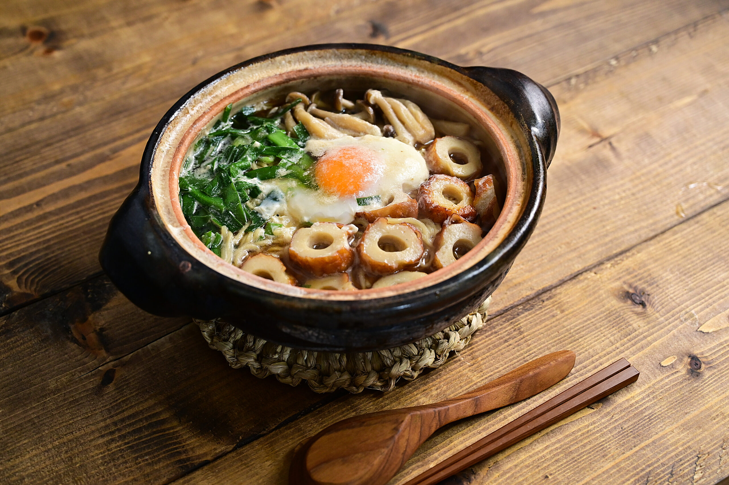 Clay pot noodle soup with greens, an egg, and fried fish cakes on a wooden table.