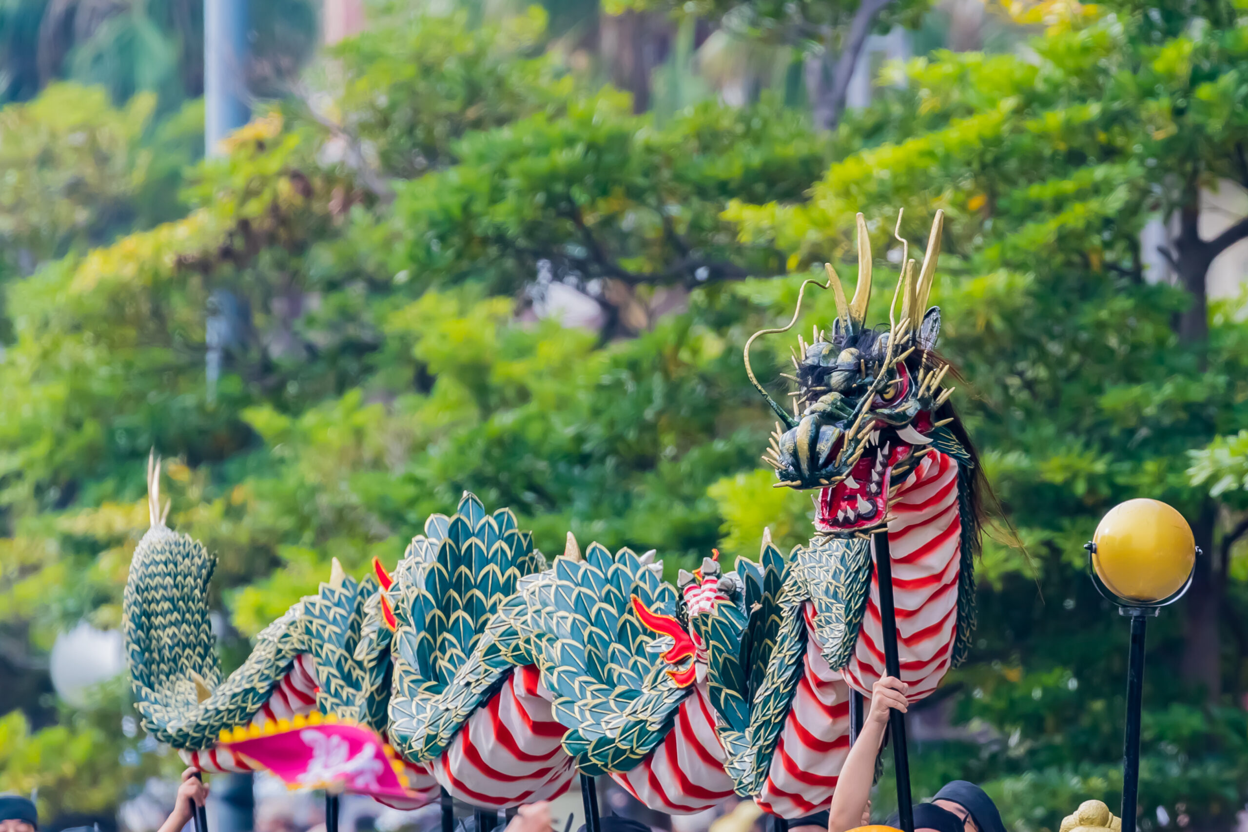 Dragon dance performer with a blue-scaled dragon costume and red-and-white stripes in a park setting.