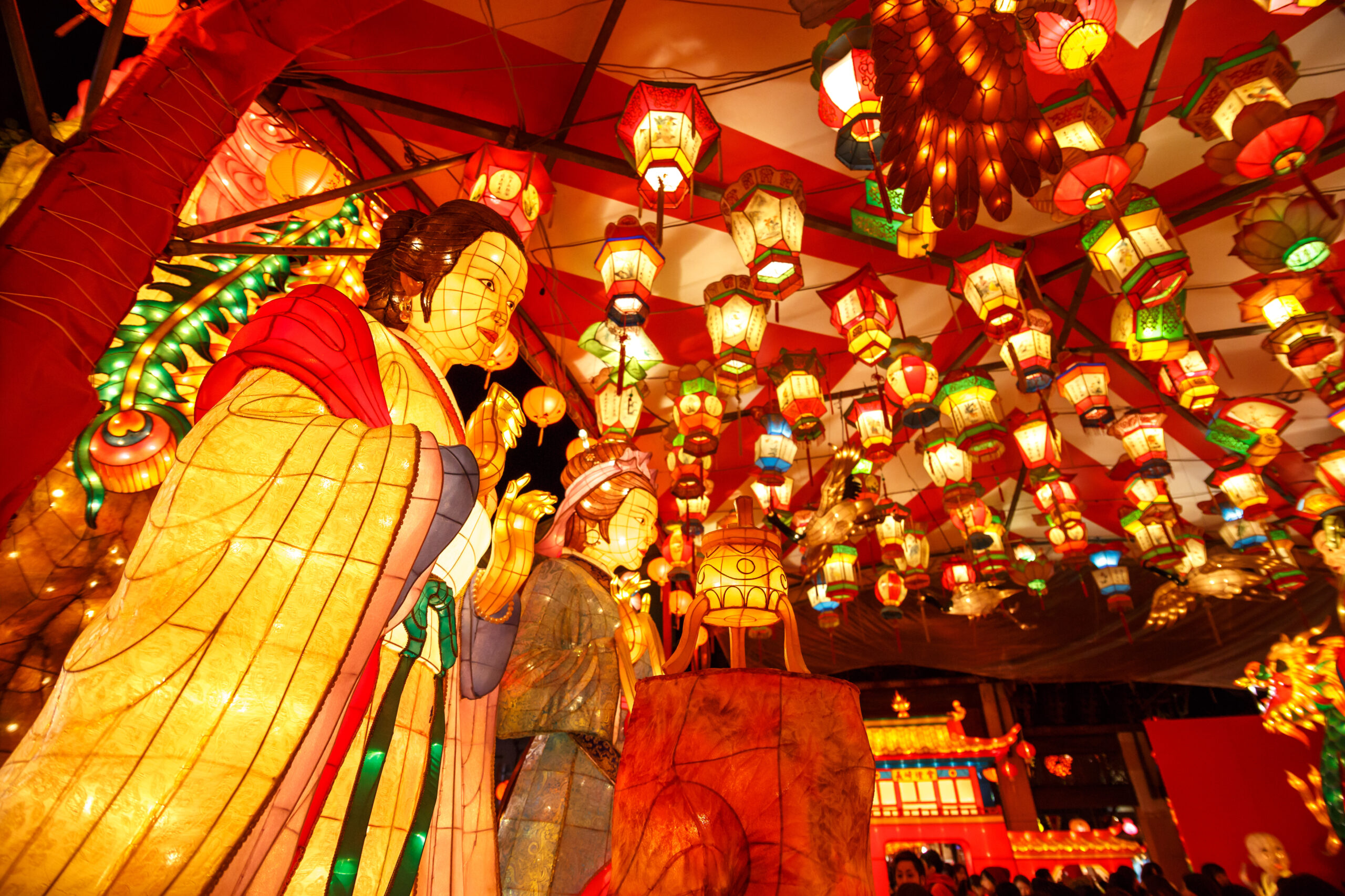 Giant illuminated statue in yellow robes beneath a ceiling of colorful hanging lanterns at a festival scene.