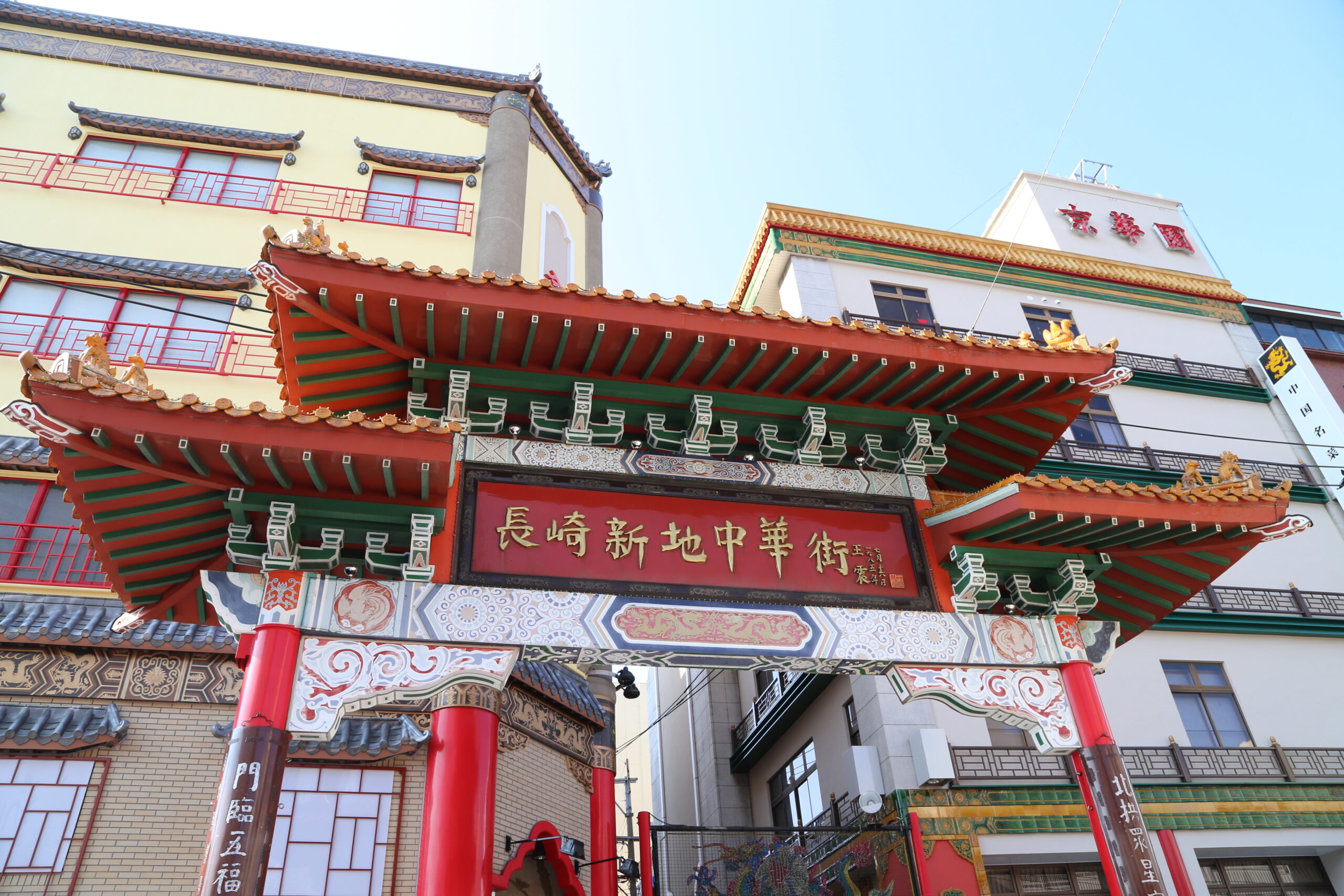 Ornate Chinese temple gate with red columns, green brackets, and gold detailing against a pale-yellow building façade and blue sky