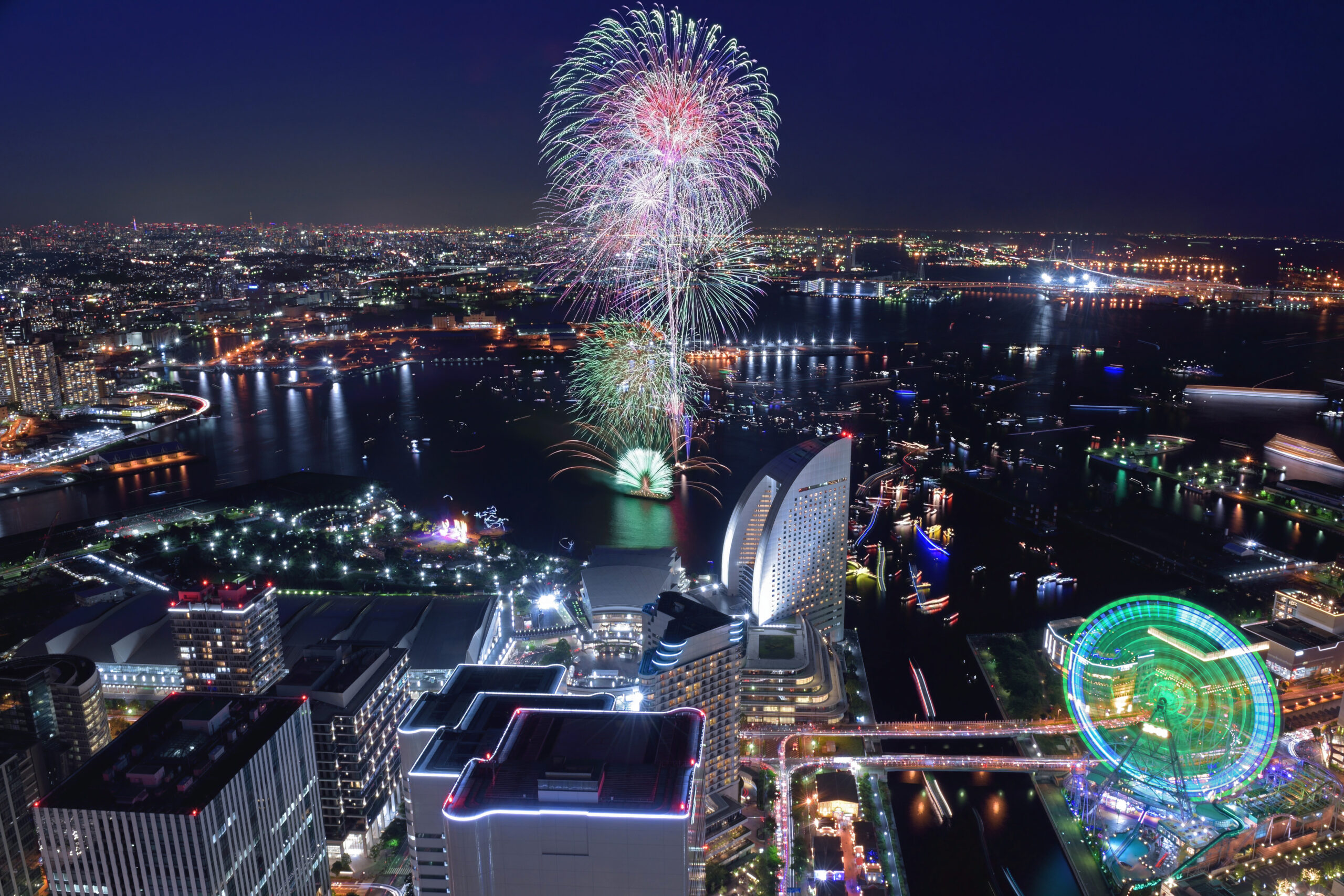 Night cityscape over a harbor with colorful fireworks lighting the sky and a tall illuminated skyline including a large Ferris wheel in the foreground.
