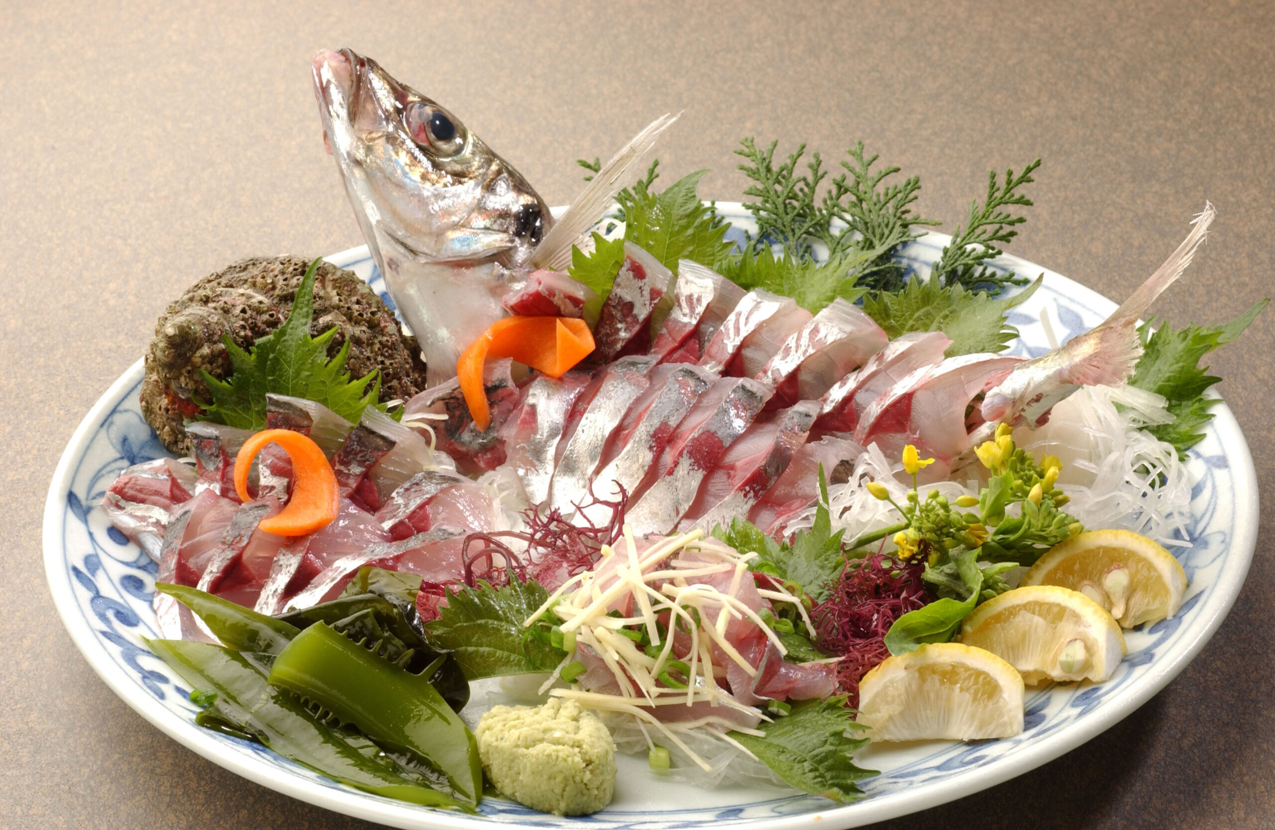 Sashimi platter with sliced fish, fresh greens, citrus wedges, wasabi, and shredded radish on a blue-and-white plate.