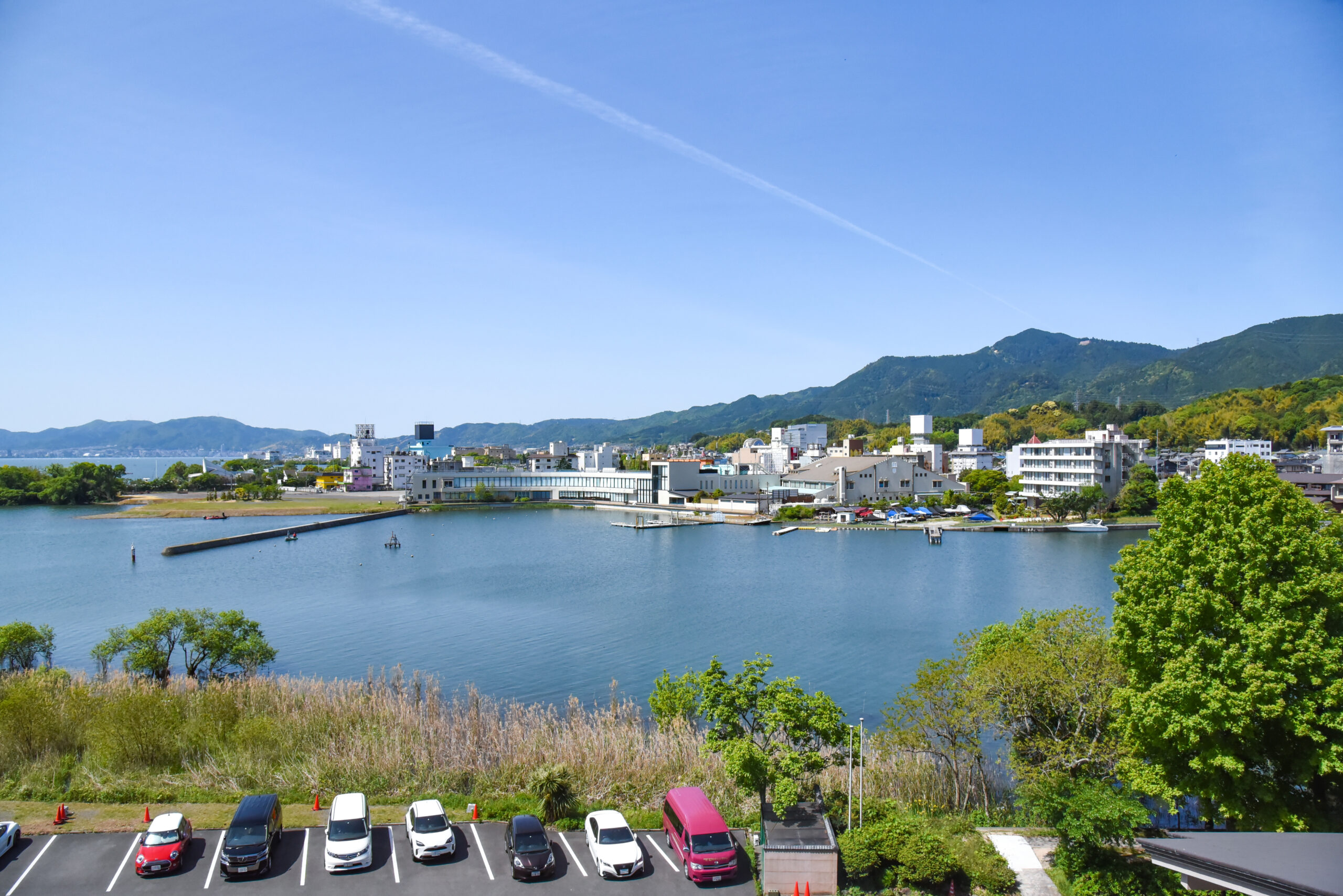 Parking lot with cars along a waterfront, buildings at the shore and green hills in the distance under a clear blue sky.