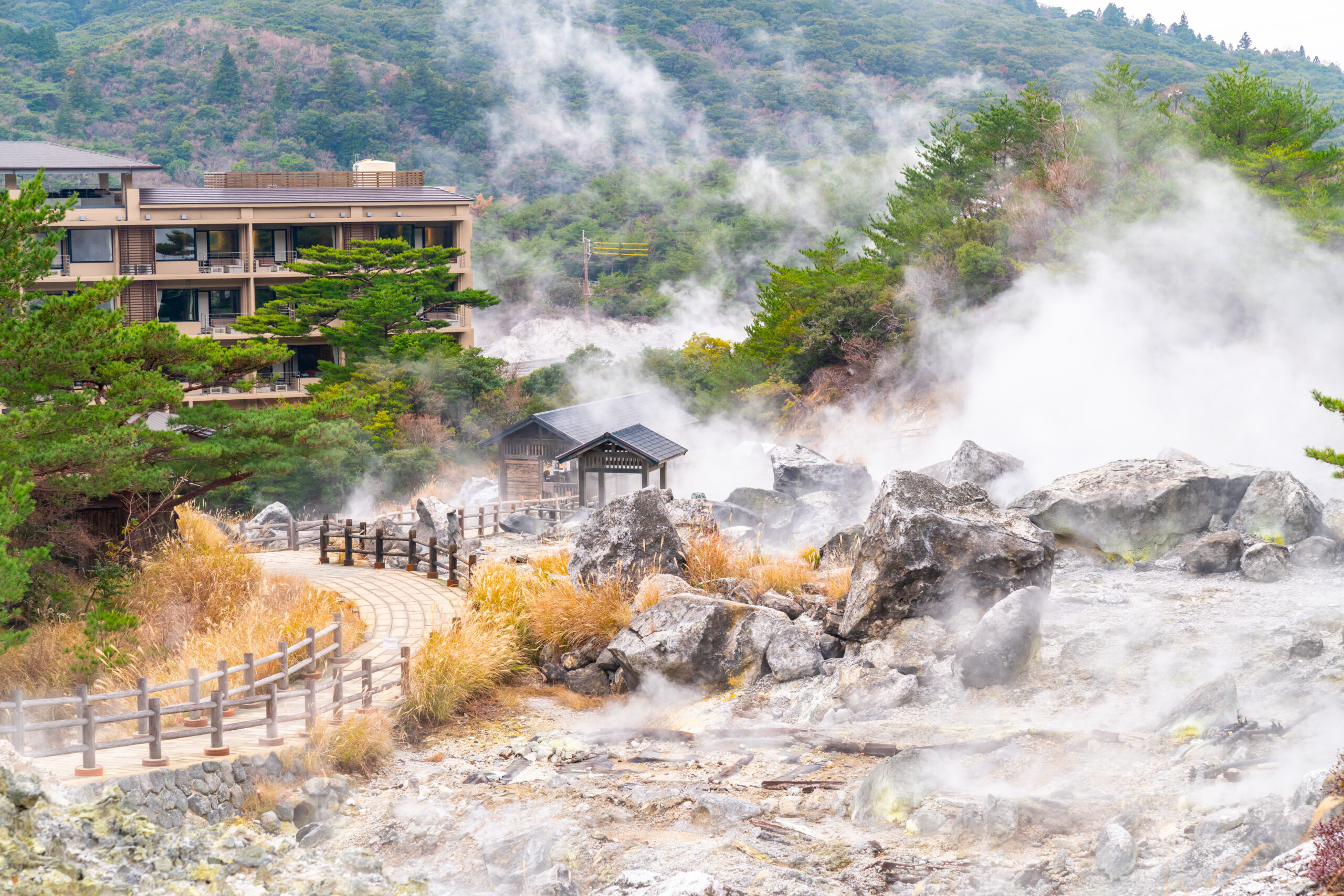 Path winds through a rocky geothermal area with steam rising, a beige hotel to the left, and forested hills in the background
