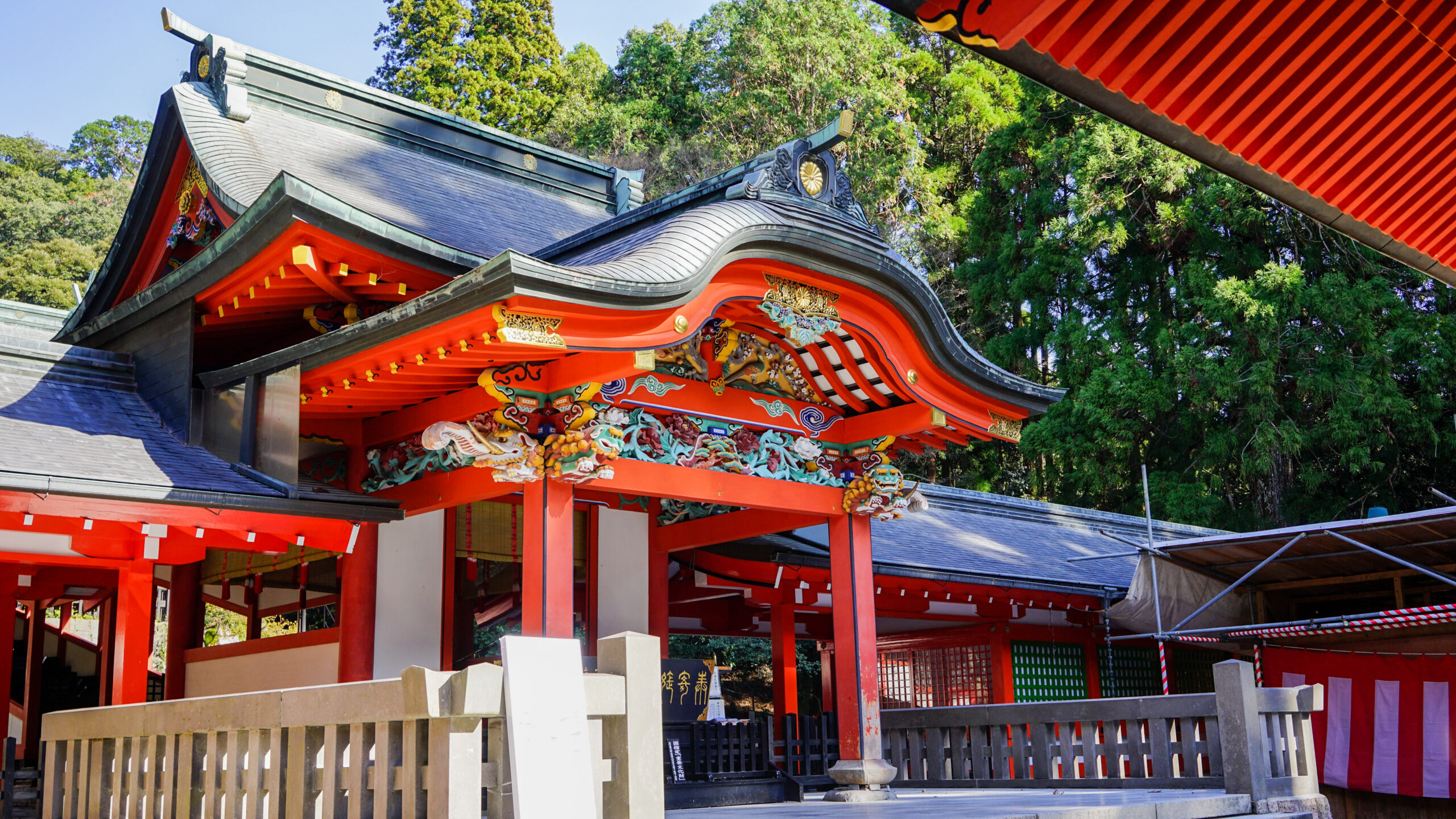 Shinto shrine roof with vivid red beams, black curved tiles, and ornate gold carvings, set against green trees.