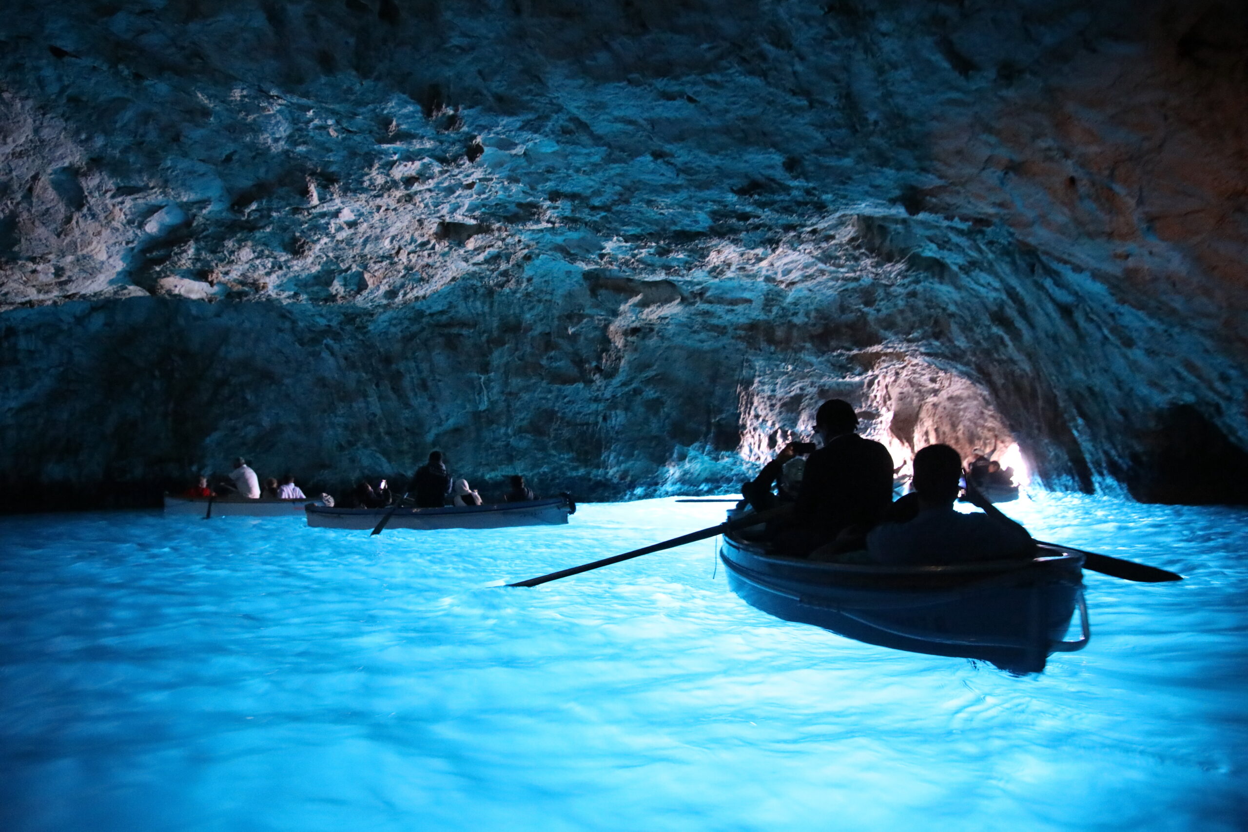 People in small boats paddling through a blue-lit cave with rocky walls and calm water reflections.