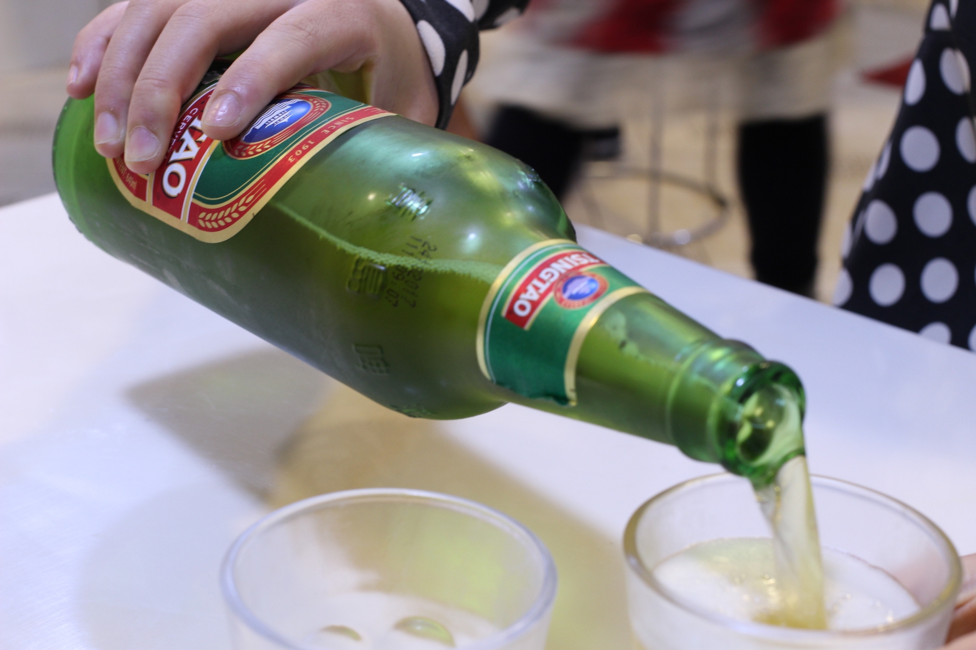 Green bottle tilted to pour sparkling wine into clear plastic cups on a white table.