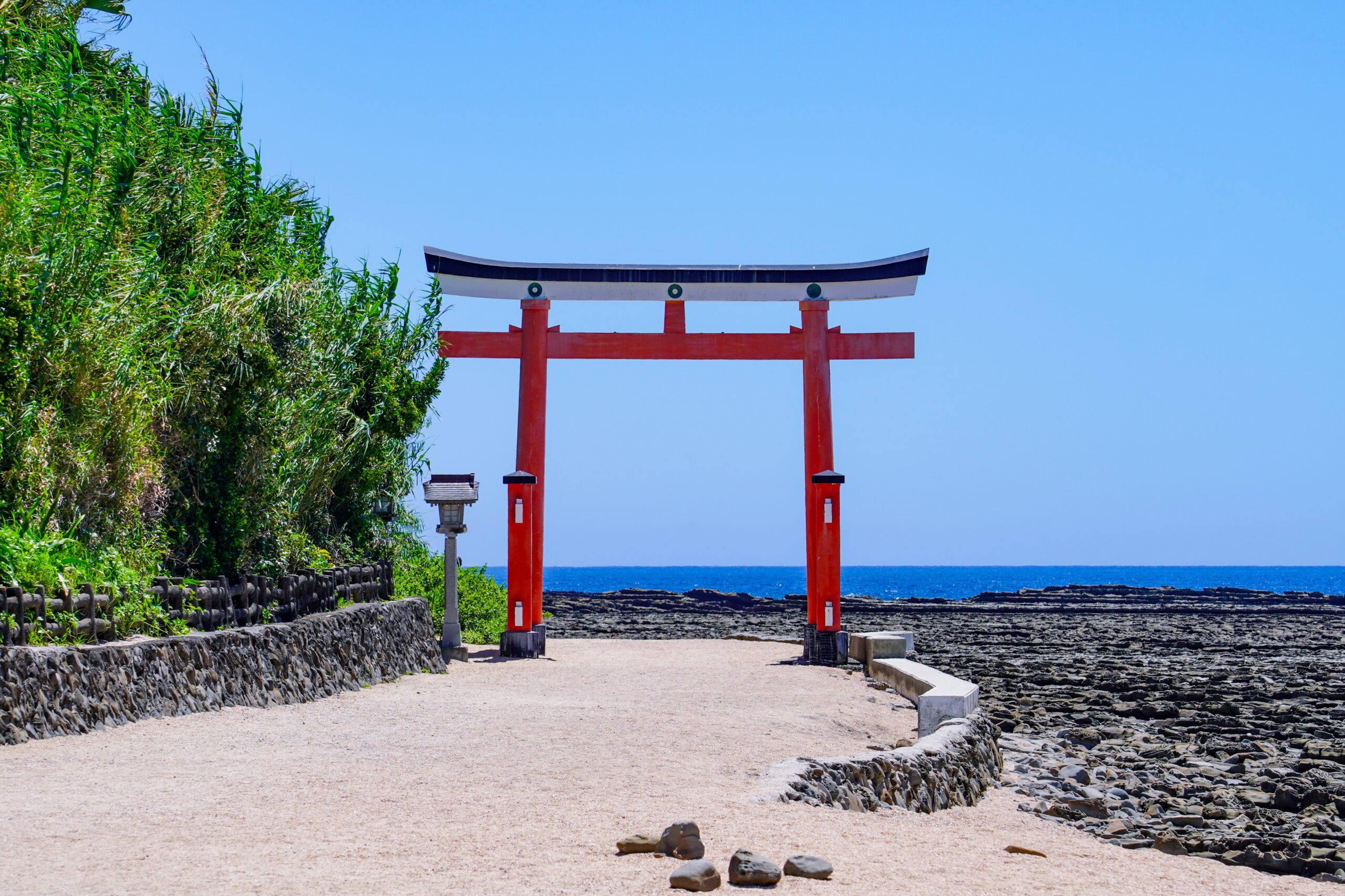 Red torii gate standing on a rocky path beside the ocean, with green foliage on the left and blue sky above.