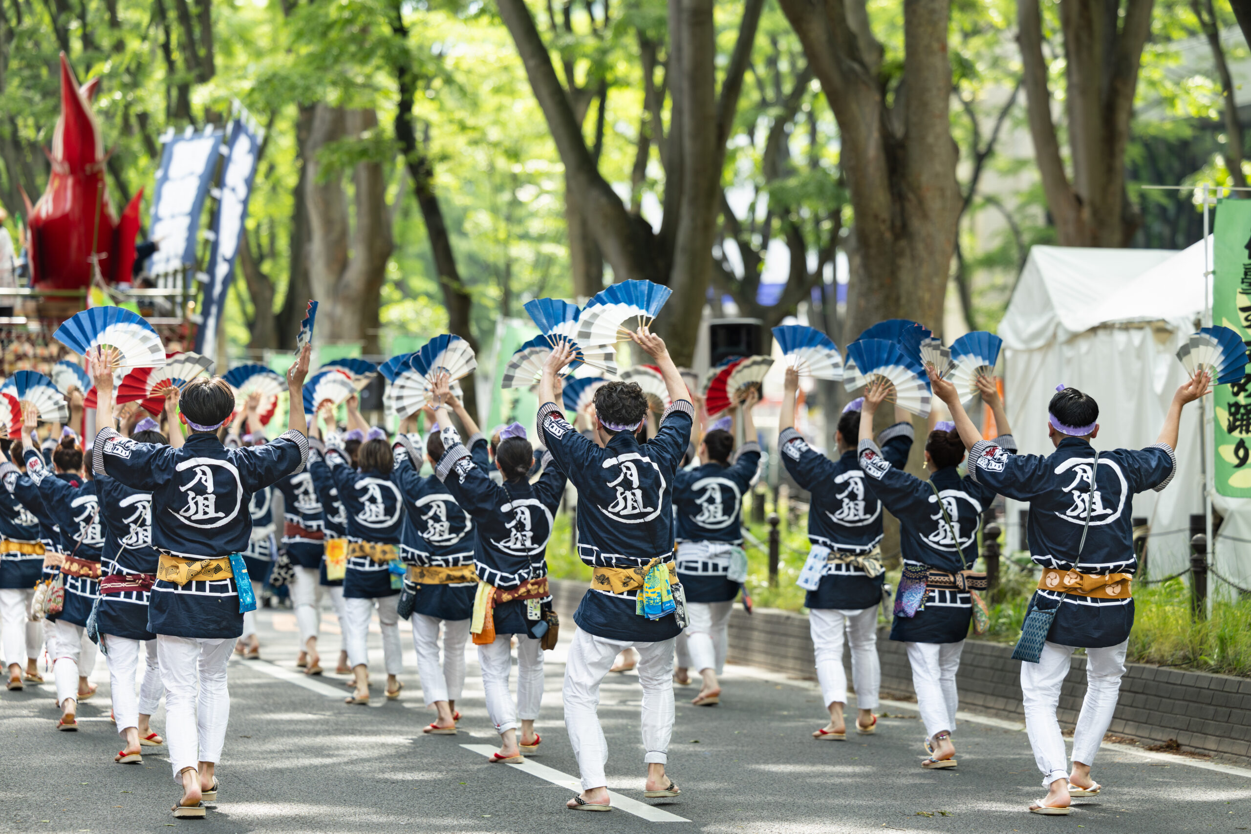 Dancers in matching blue jackets and white pants parade down a city street, waving blue-and-white fans overhead.