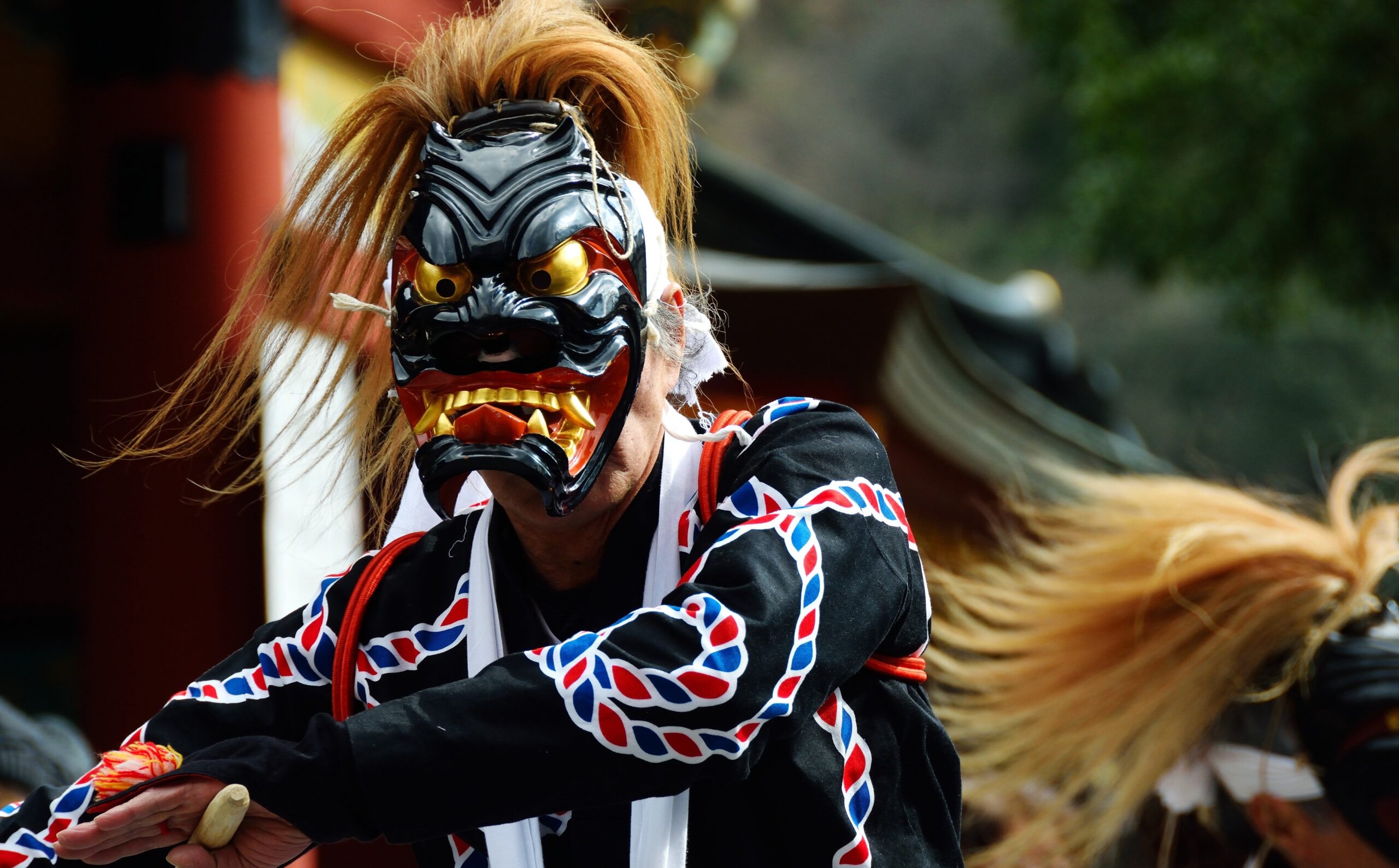 Person wearing a black and red Oni mask with gold eyes, performing a traditional dance festival show.
