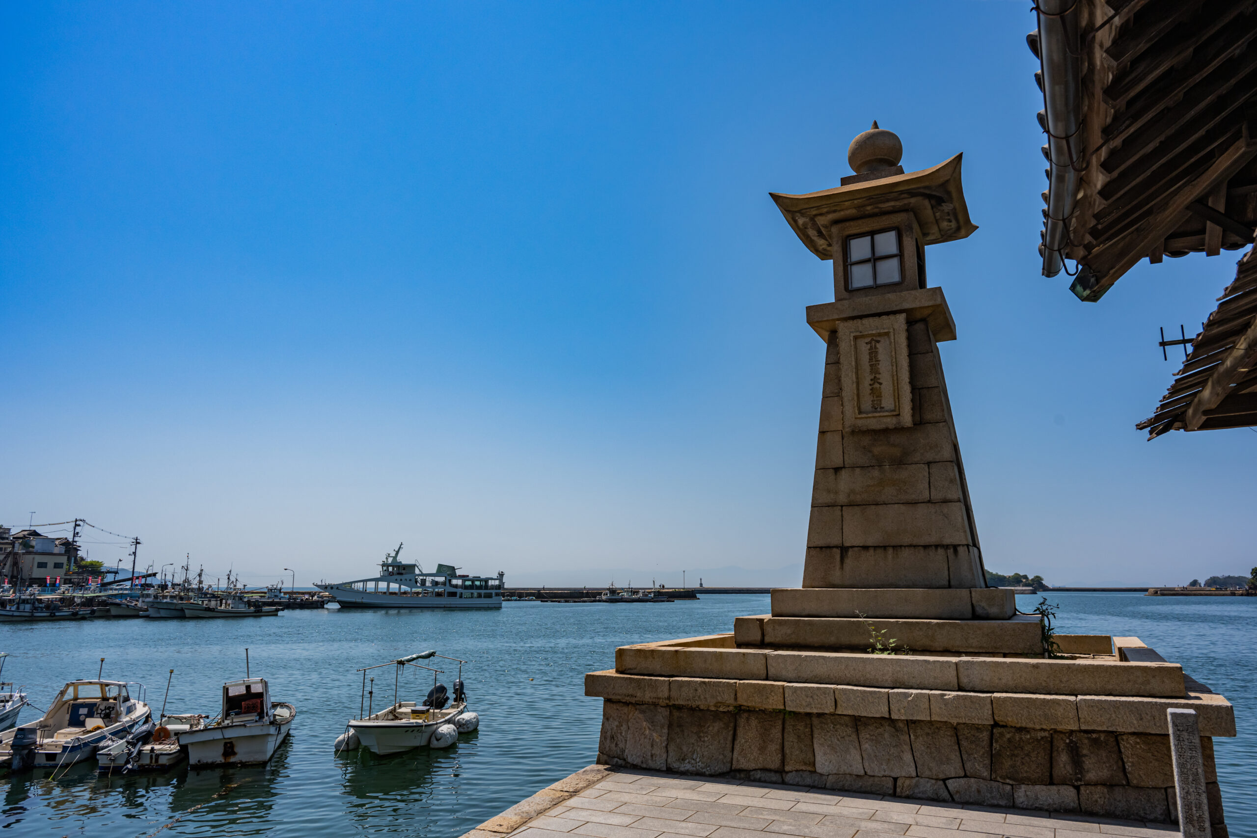 Harbor scene with small boats docked along calm water and a tall stone lantern on the right under a clear blue sky.