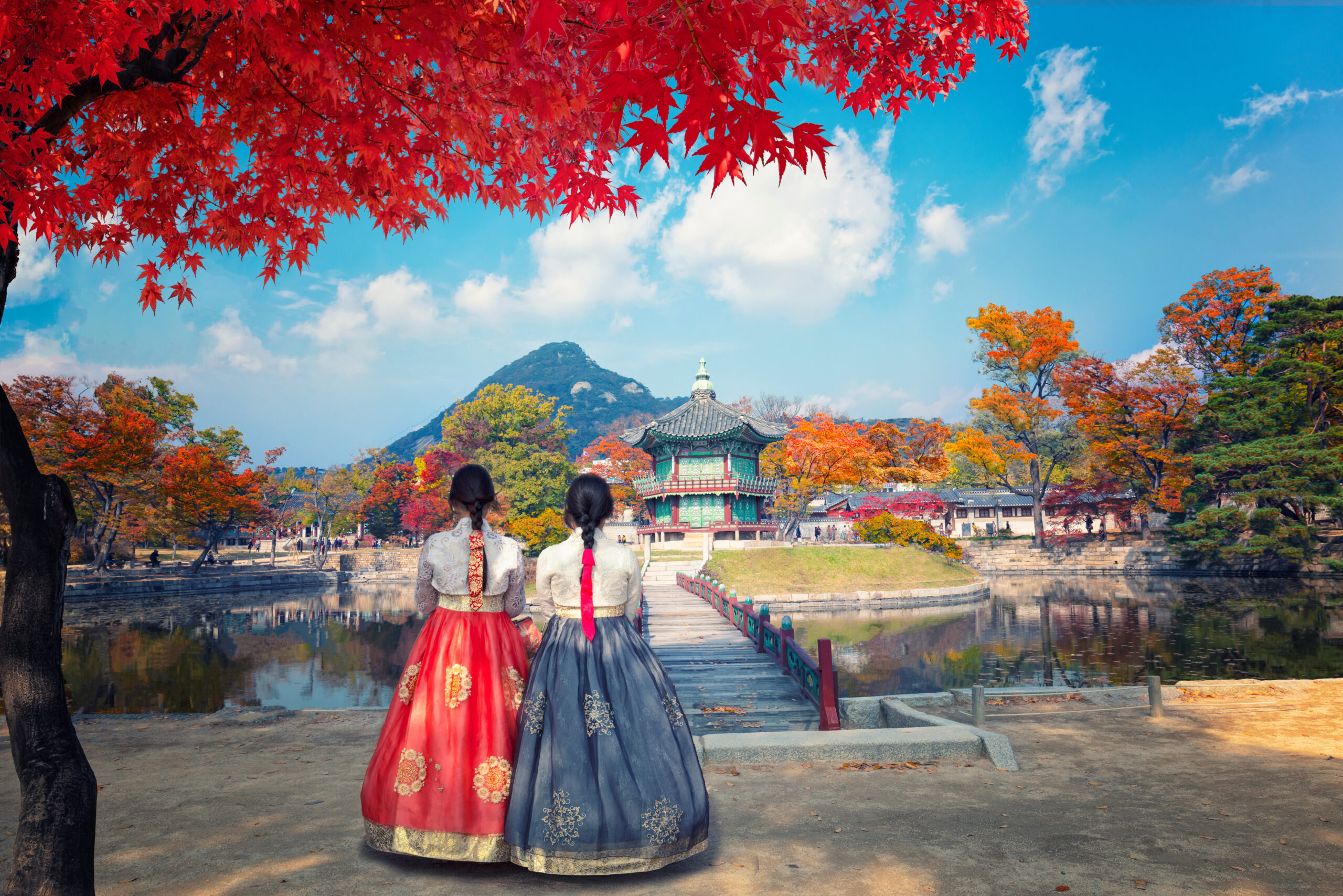 Back view of two women in vibrant hanbok standing on a wooden bridge in a park with red autumn leaves overhead and a pagoda in the distance, mountains beyond
