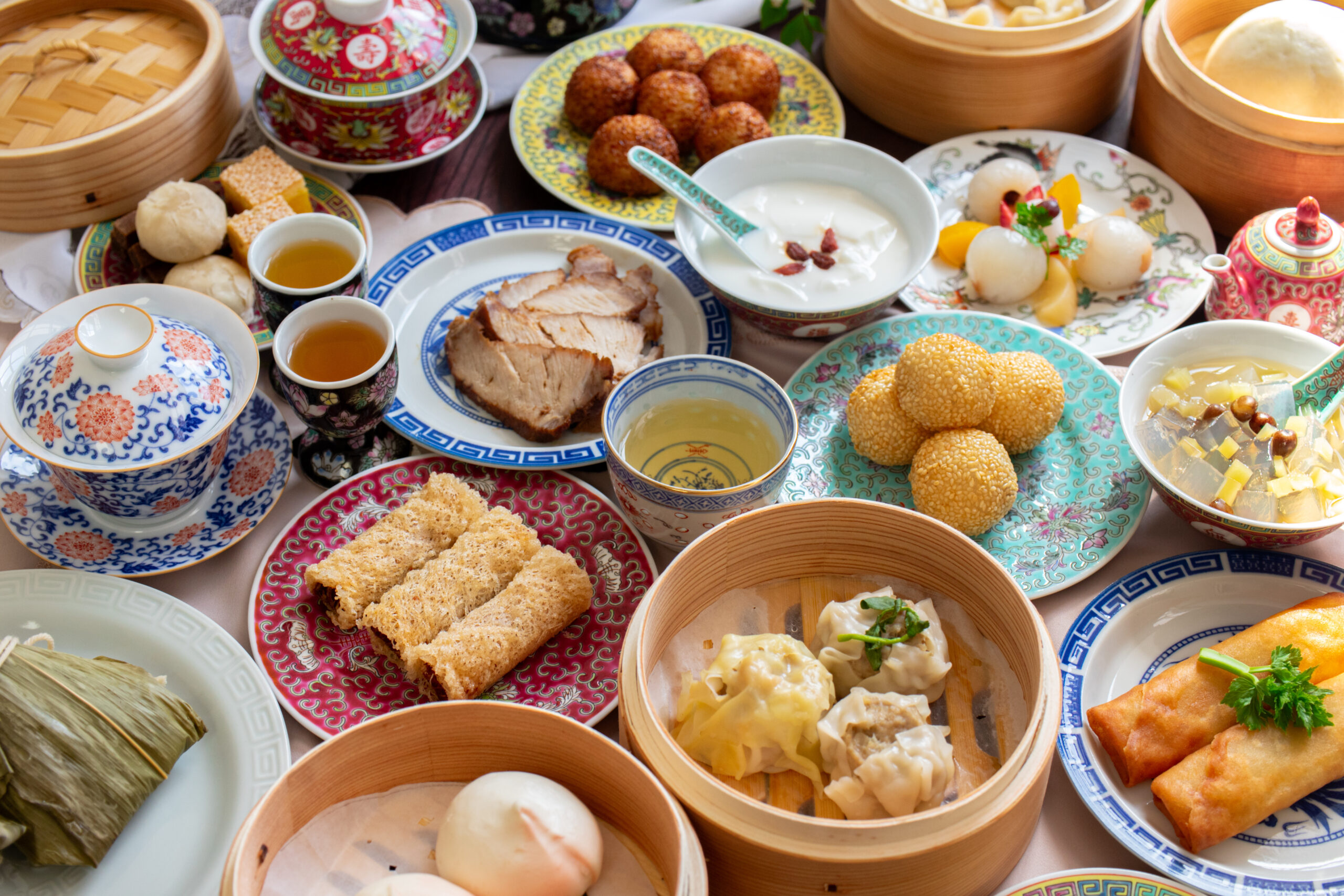 A colorful spread of dim sum and Chinese dishes on decorative plates and bamboo steamers, with dumplings, buns, and fried snacks arranged together.