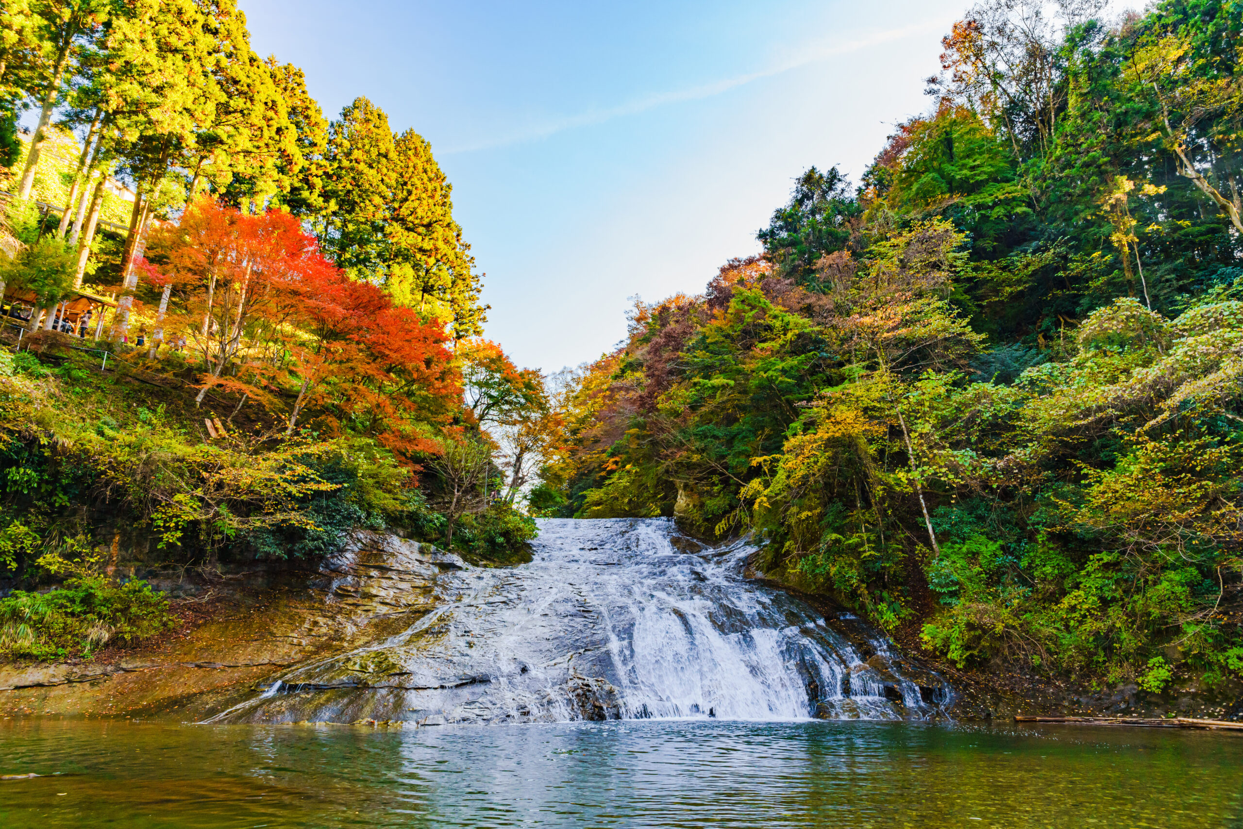 Autumn forest surrounds a multi-tiered waterfall flowing into a calm pool below.