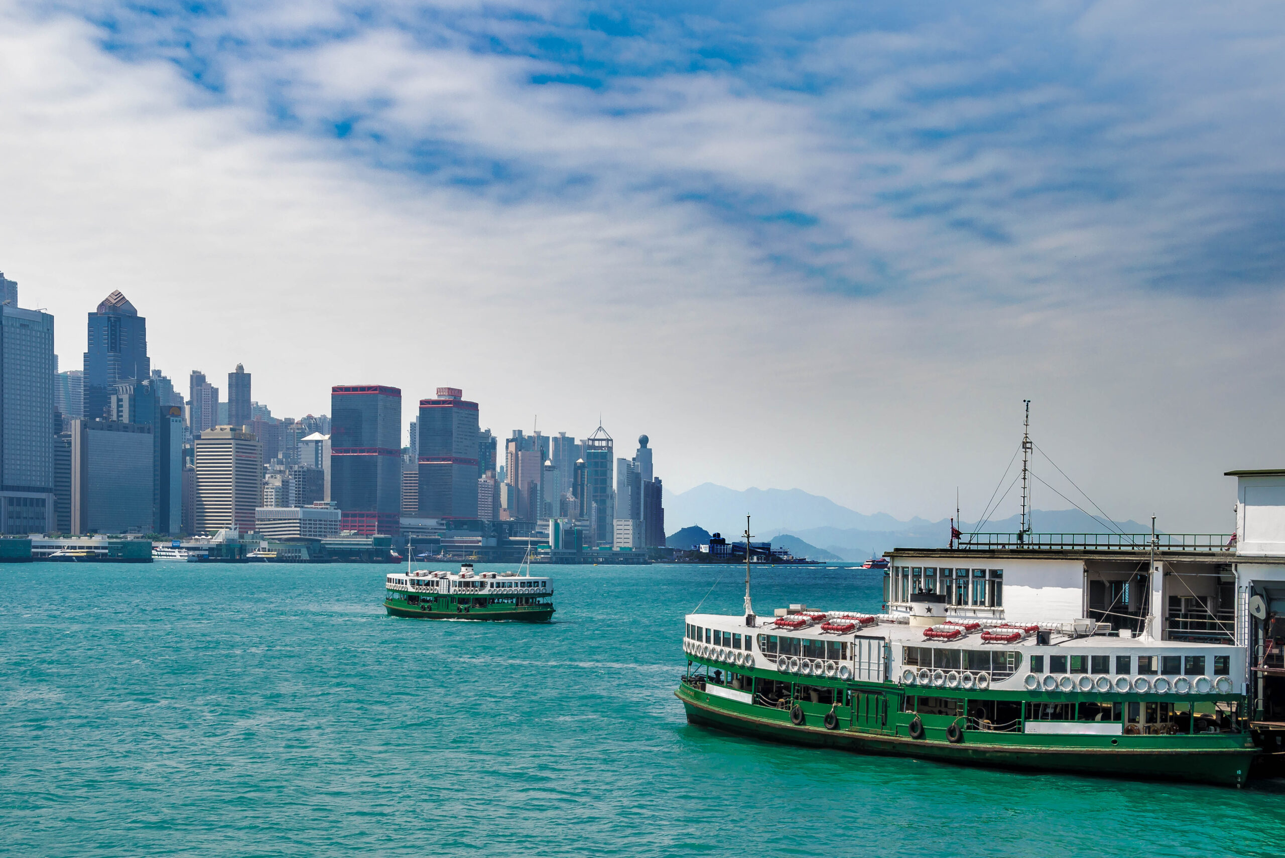 City skyline along a turquoise harbor with a large green-and-white passenger ferry in the foreground under a blue, cloudy sky.