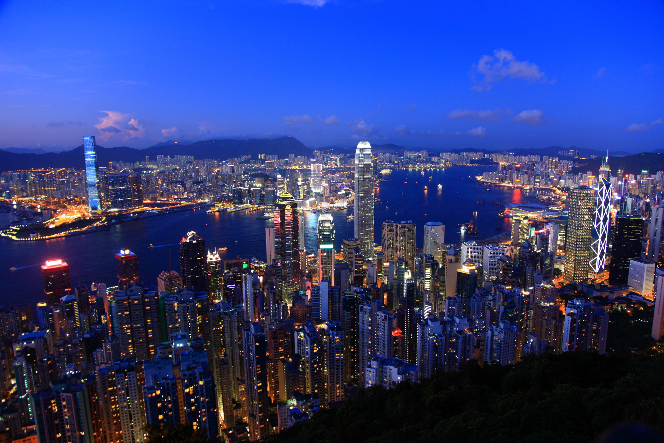 Nighttime aerial view of Hong Kong's illuminated skyline along Victoria Harbour, with tall skyscrapers and distant mountains.