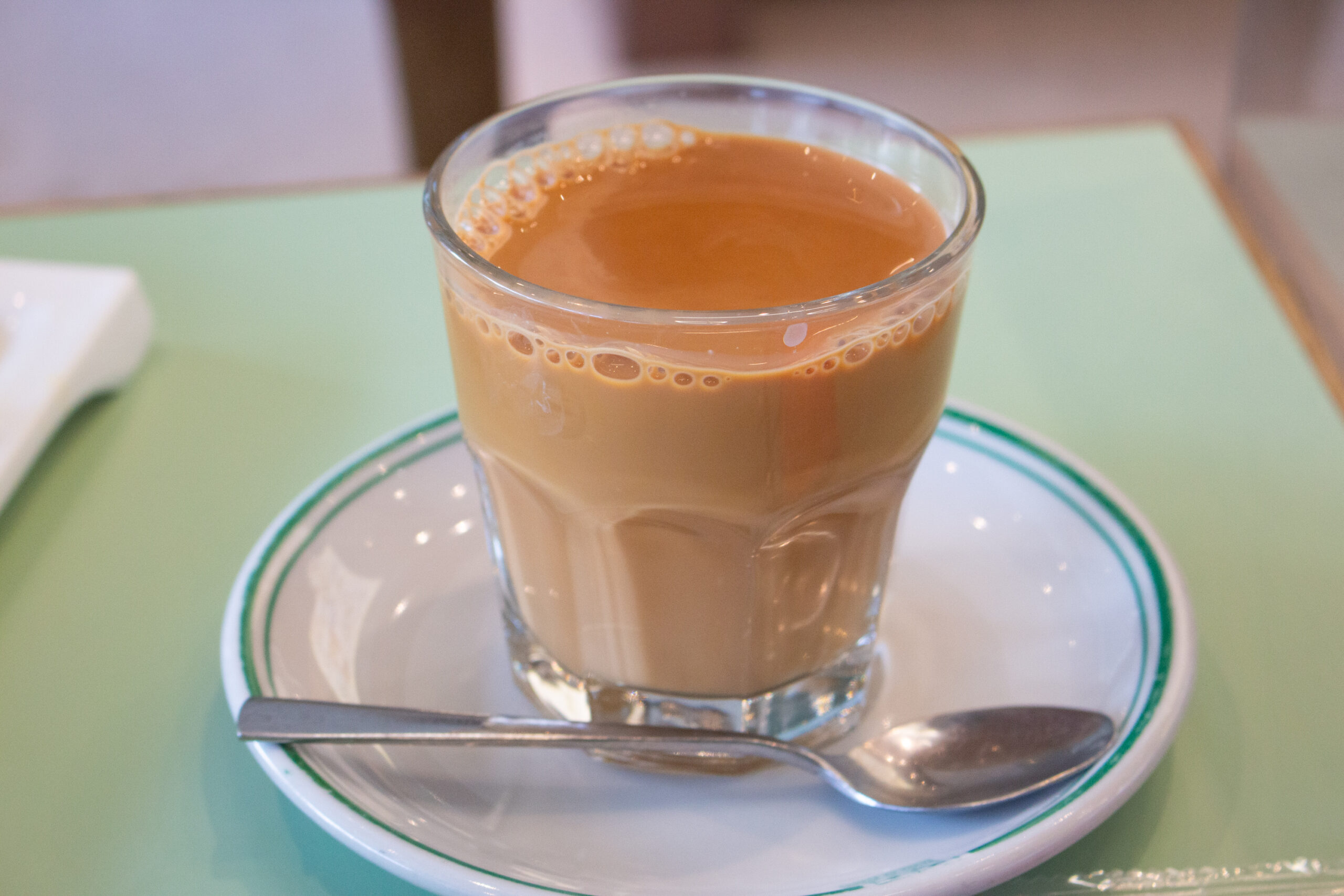 Tall glass of iced or warm coffee with creamy light brown color on a white saucer with green trim and a spoon laid across the plate