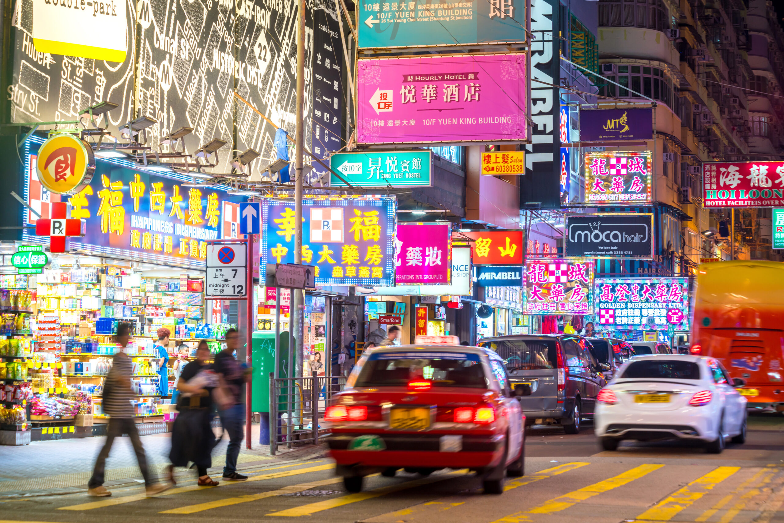 Bustling neon-lit street in Hong Kong with colorful storefront signs, pedestrians, and traffic at dusk.