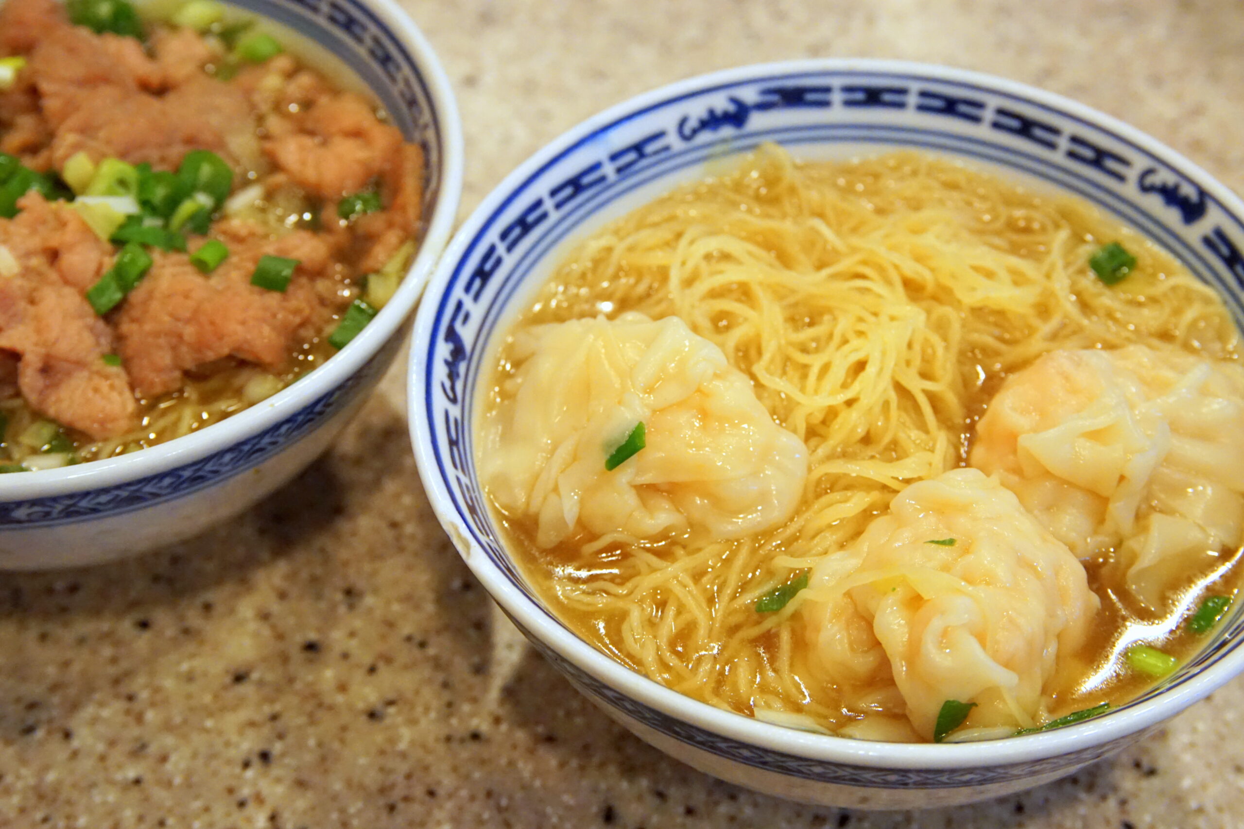 Two bowls of Asian dishes: noodle dumpling soup in a blue-patterned bowl next to a fried meat dish with scallions, on a speckled countertop.