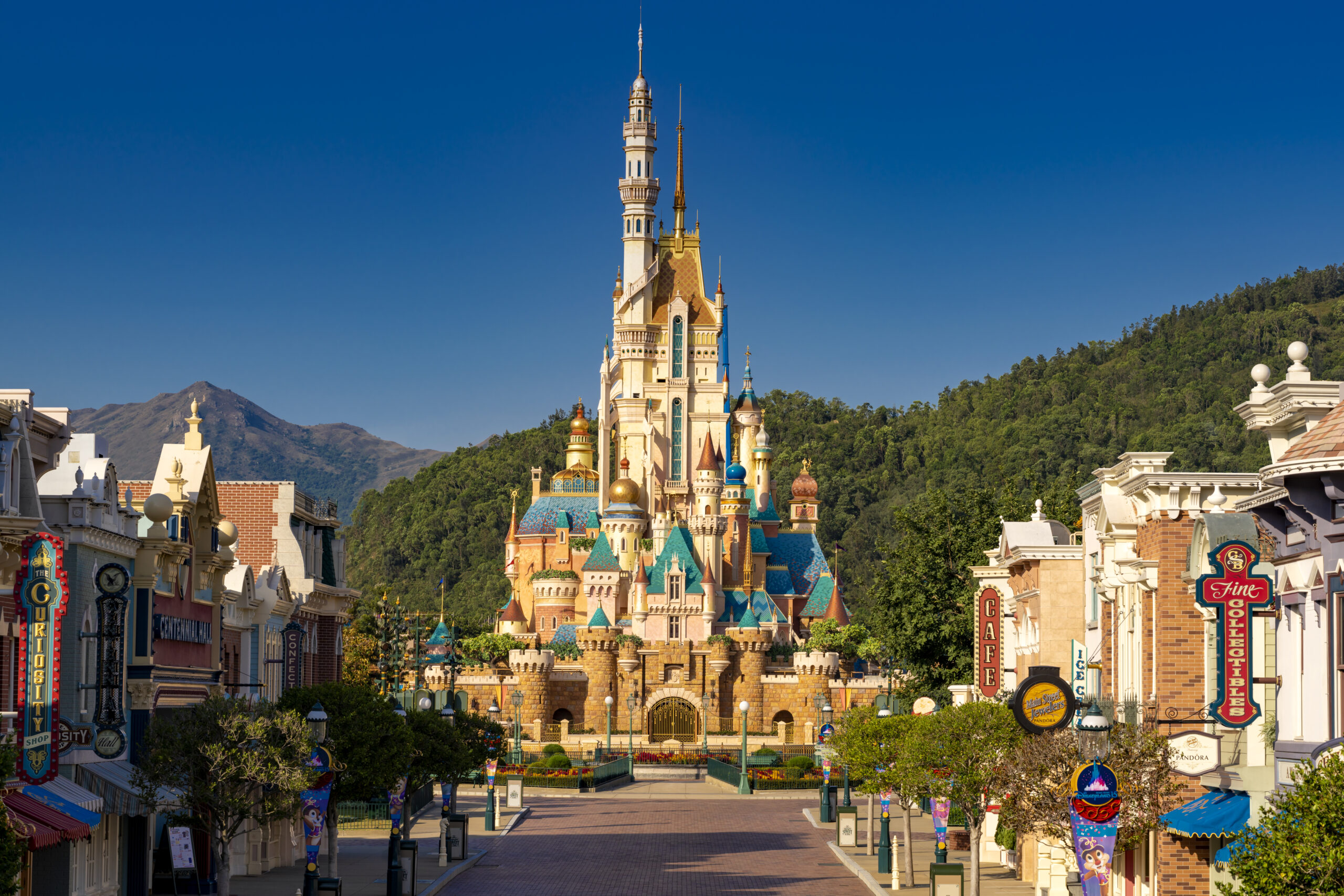 Disney-like castle central to an amusement park, with colorful towers and storefronts lining a wide pedestrian street against a blue sky