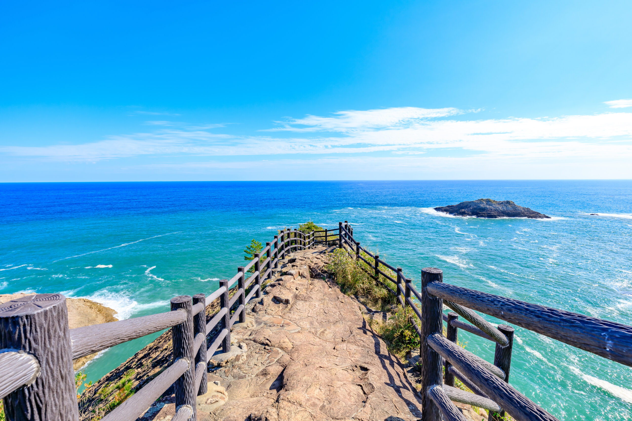 Winding rocky path with weathered wooden railing along a cliff, leading to the turquoise ocean and a small rocky island under a clear blue sky.